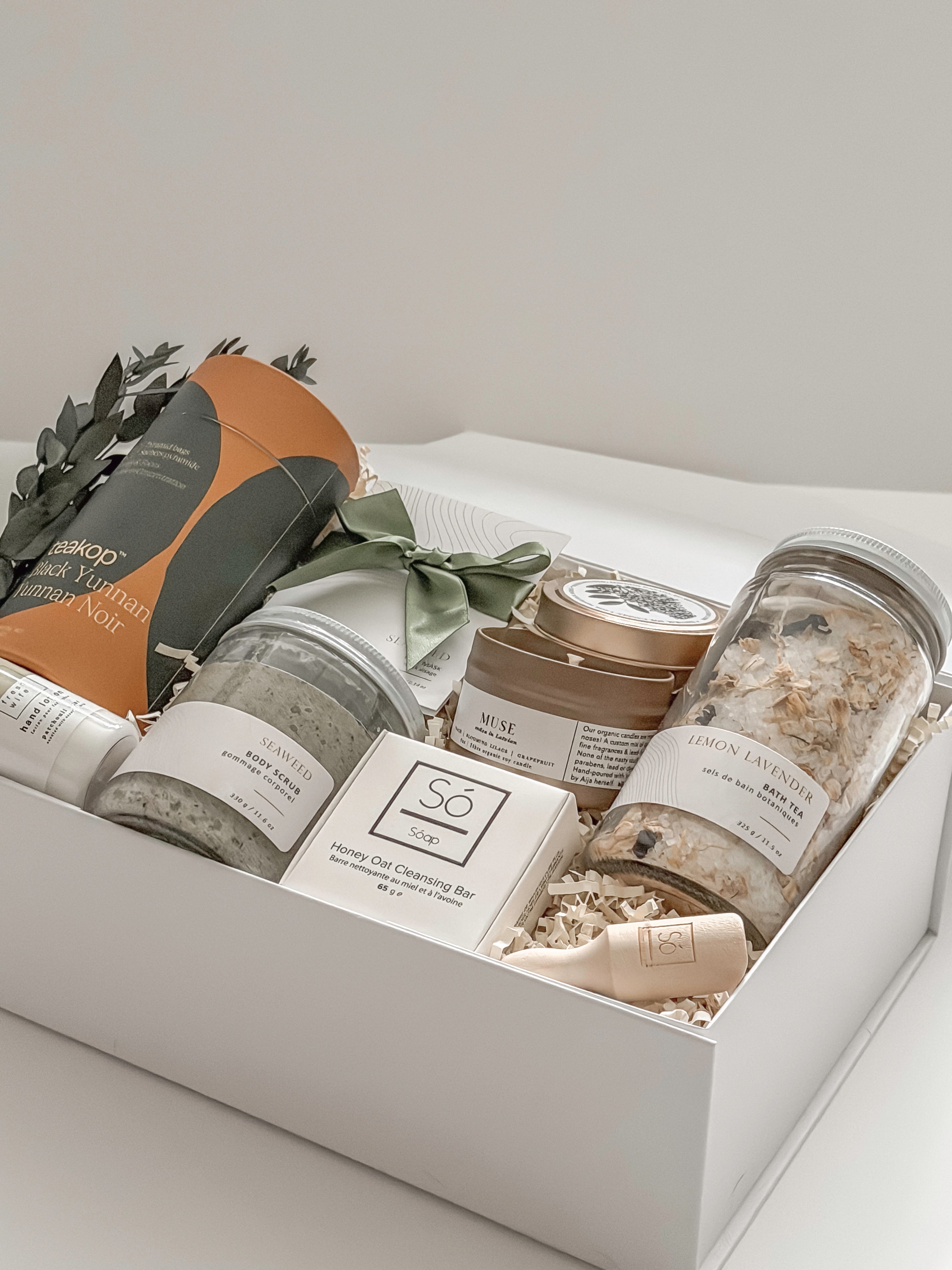 Élysée spa gift box by Muse Baskets and Co. shown from above, featuring Teakop Black Yunnan tea, a Muse candle by Aija, Sealuxe seaweed body scrub and face mask and lemon lavender bath tea, a So Luxury honey oat cleansing bar and wooden scoop, and patchouli mint hand lotion in a white box with eucalyptus and olive green ribbon, a refined thank you, employee appreciation, and client appreciation gift.