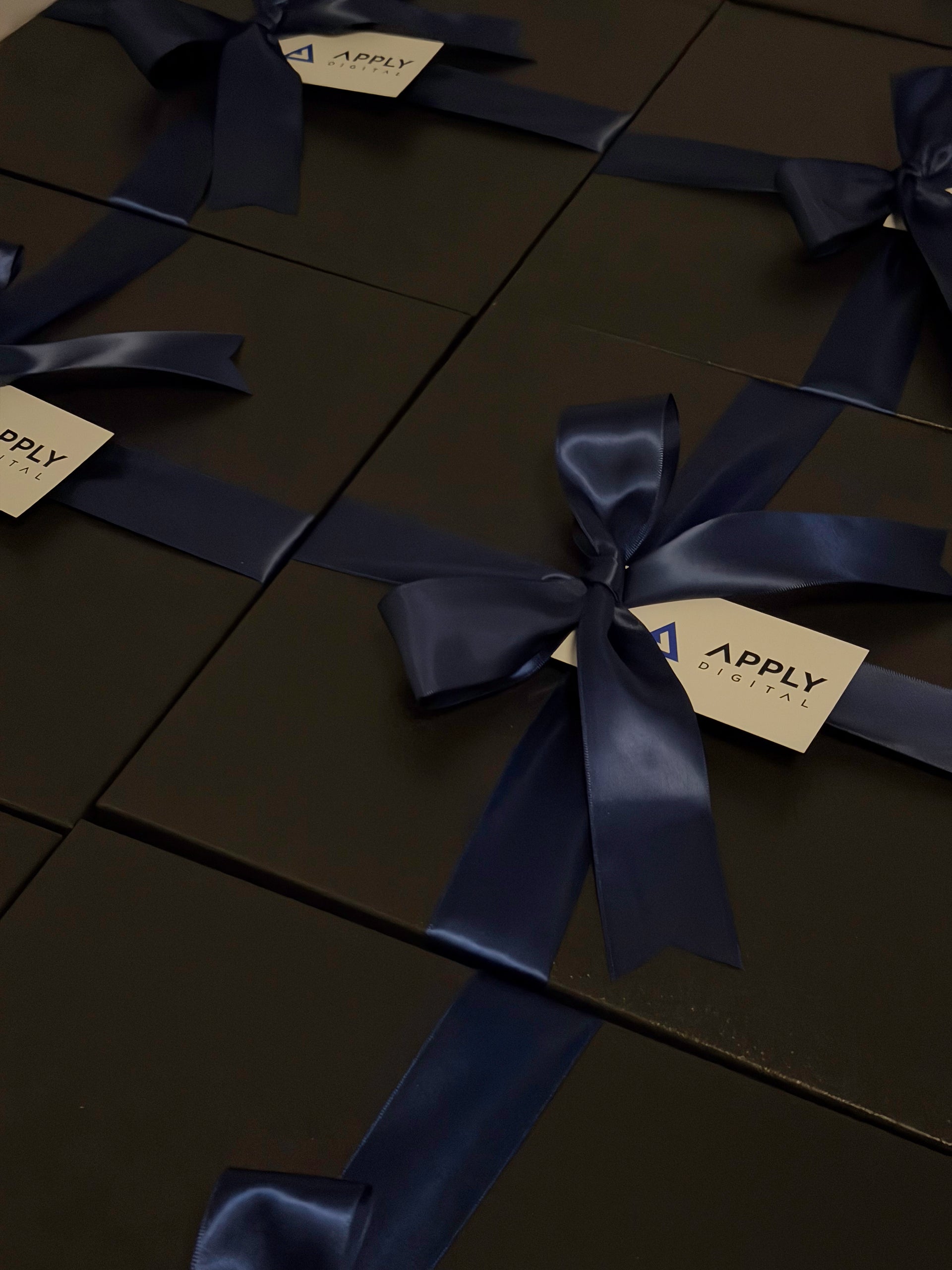 Dark charcoal gift boxes tied with navy blue satin ribbons and bow, each with a small white tag printed with the brand name 'APPLY'.