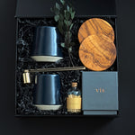 The Lennox gift box by Muse Baskets and Co. viewed close up at an angle, showing two midnight blue Fable mugs, two olive wood coasters, a VIS myrrh, amber, and sandalwood candle by Vancouver Candle Co., a gold candle snuffer, and black tipped matches in an apothecary jar with eucalyptus in a black box, a refined possession day, homeowner welcome, and development possession gift.