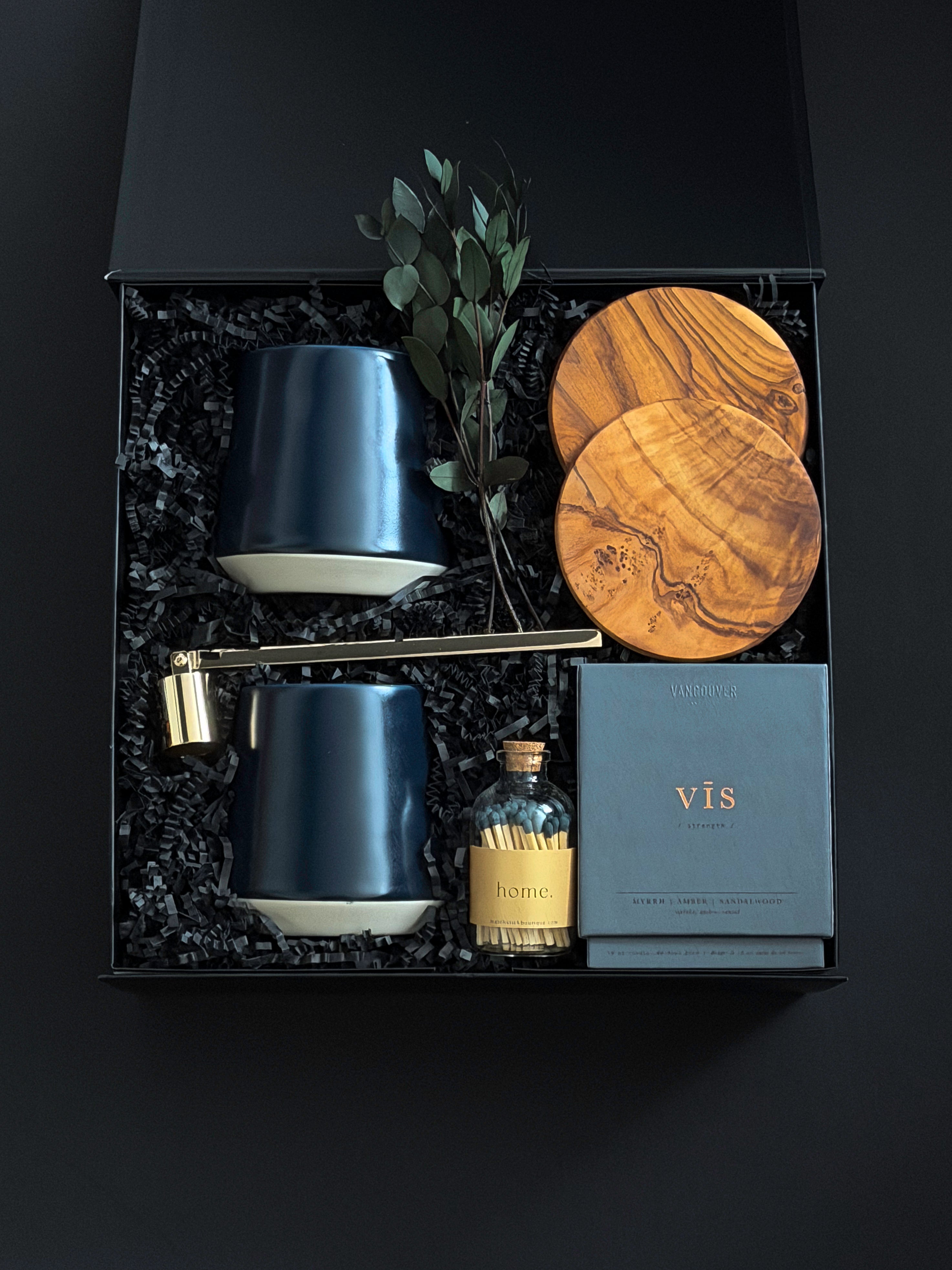 The Lennox gift box by Muse Baskets and Co. viewed close up at an angle, showing two midnight blue Fable mugs, two olive wood coasters, a VIS myrrh, amber, and sandalwood candle by Vancouver Candle Co., a gold candle snuffer, and black tipped matches in an apothecary jar with eucalyptus in a black box, a refined possession day, homeowner welcome, and development possession gift.