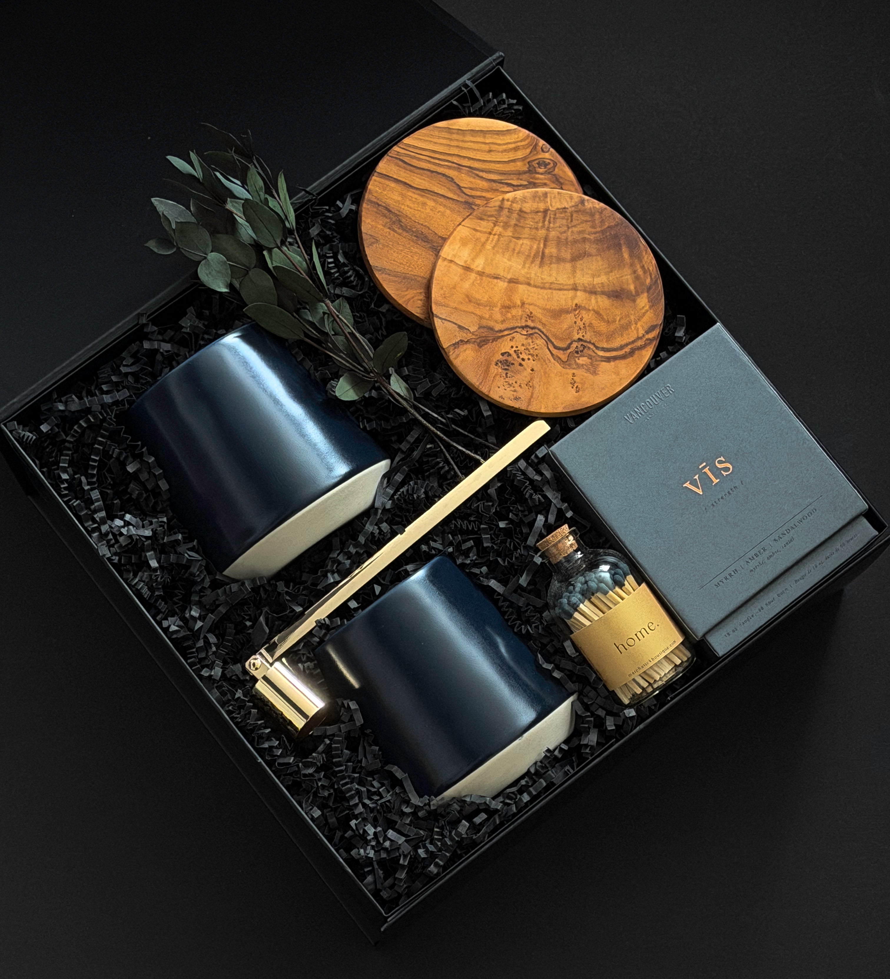 The Lennox gift box by Muse Baskets and Co. shown from above, featuring two midnight blue Fable mugs, two olive wood coasters, a VIS candle by Vancouver Candle Co., a gold candle snuffer, and black tipped matches in an apothecary jar with eucalyptus on black fill in a black box, a refined realtor closing gift, possession day gift, and homeowner welcome gift.