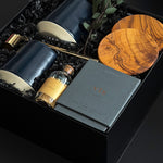 The Lennox gift box by Muse Baskets and Co. photographed at an angle, with two midnight blue Fable mugs, olive wood coasters, a VIS candle by Vancouver Candle Co., a gold snuffer, and black tipped matches in an apothecary jar nestled in a black box with eucalyptus, a curated realtor, developer, and corporate closing gift from Canadian makers.
