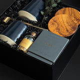 The Lennox gift box by Muse Baskets and Co. photographed at an angle, with two midnight blue Fable mugs, olive wood coasters, a VIS candle by Vancouver Candle Co., a gold snuffer, and black tipped matches in an apothecary jar nestled in a black box with eucalyptus, a curated realtor, developer, and corporate closing gift from Canadian makers.