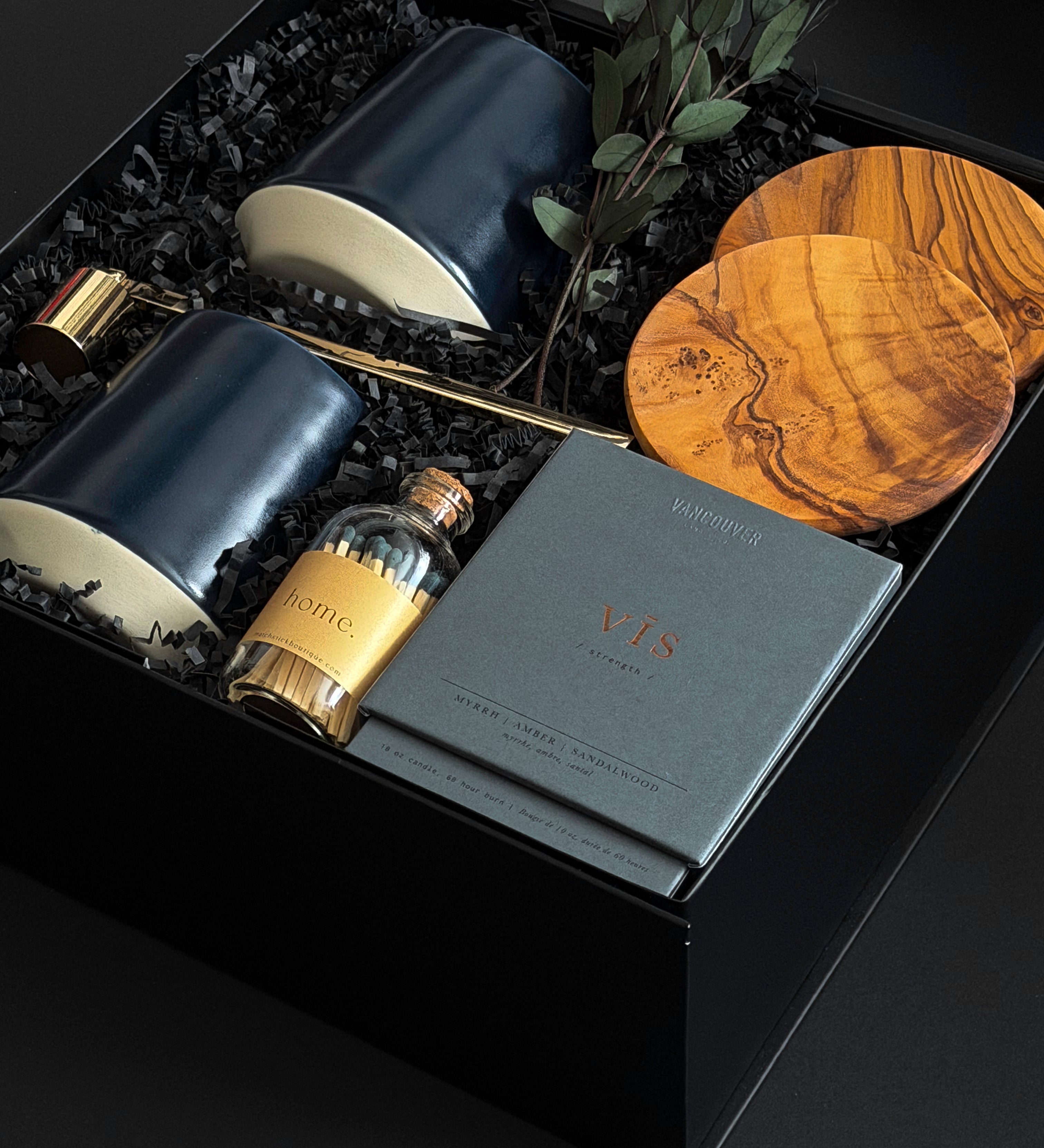 The Lennox gift box by Muse Baskets and Co. photographed at an angle, with two midnight blue Fable mugs, olive wood coasters, a VIS candle by Vancouver Candle Co., a gold snuffer, and black tipped matches in an apothecary jar nestled in a black box with eucalyptus, a curated realtor, developer, and corporate closing gift from Canadian makers.