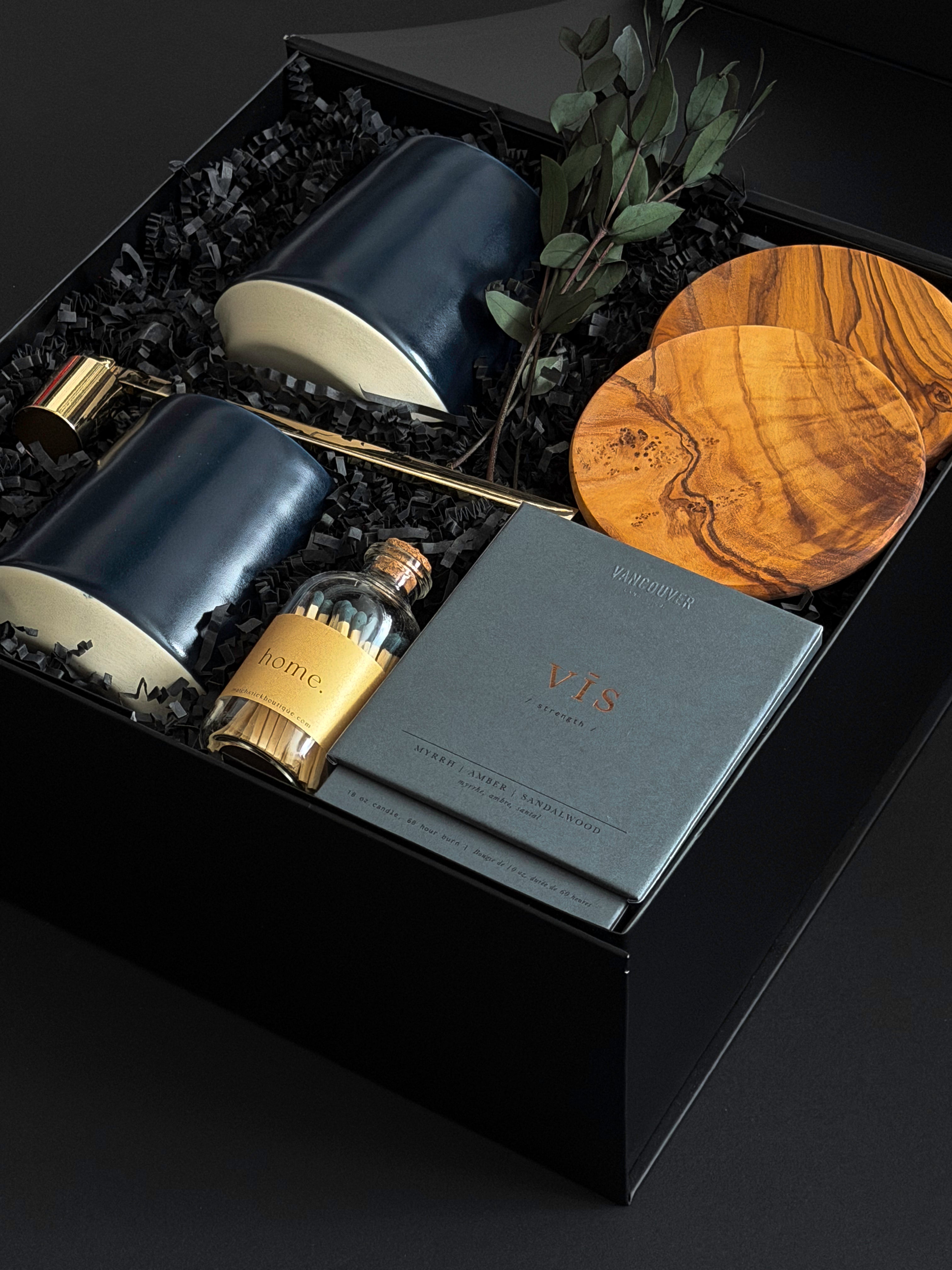 The Lennox gift box by Muse Baskets and Co. photographed at an angle, with two midnight blue Fable mugs, olive wood coasters, a VIS candle by Vancouver Candle Co., a gold snuffer, and black tipped matches in an apothecary jar nestled in a black box with eucalyptus, a curated realtor, developer, and corporate closing gift from Canadian makers.