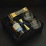 The Belgravia luxury whisky gift basket by Muse Baskets and Co. featuring The Glenlivet Founder's Reserve 1824 Single Malt Scotch Whisky, Viski Meridian gold-rimmed crystal lowball tumblers, a gold shot jigger tied with a navy blue satin ribbon, and Roger's Chocolates Dark Chocolate Champagne Truffles, presented in a signature black gift box with black crinkle fill, Vancouver, BC.