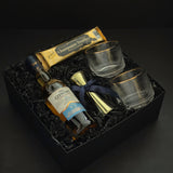 The Belgravia luxury whisky gift basket by Muse Baskets and Co. featuring The Glenlivet Founder's Reserve 1824 Single Malt Scotch Whisky, Viski Meridian gold-rimmed crystal lowball tumblers, a gold shot jigger tied with a navy blue satin ribbon, and Roger's Chocolates Dark Chocolate Champagne Truffles, presented in a signature black gift box with black crinkle fill, Vancouver, BC.