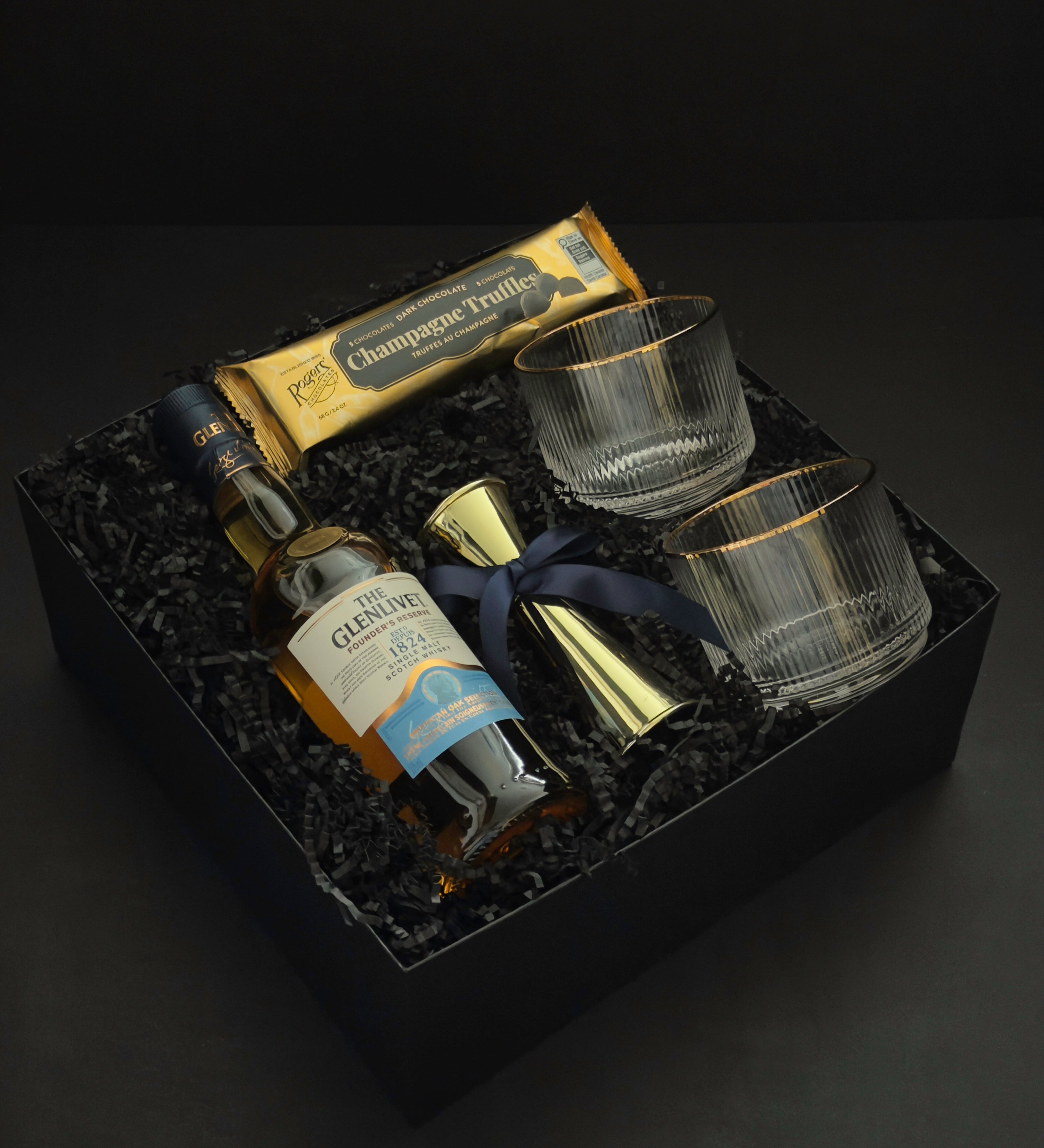 The Belgravia luxury whisky gift basket by Muse Baskets and Co. featuring The Glenlivet Founder's Reserve 1824 Single Malt Scotch Whisky, Viski Meridian gold-rimmed crystal lowball tumblers, a gold shot jigger tied with a navy blue satin ribbon, and Roger's Chocolates Dark Chocolate Champagne Truffles, presented in a signature black gift box with black crinkle fill, Vancouver, BC.