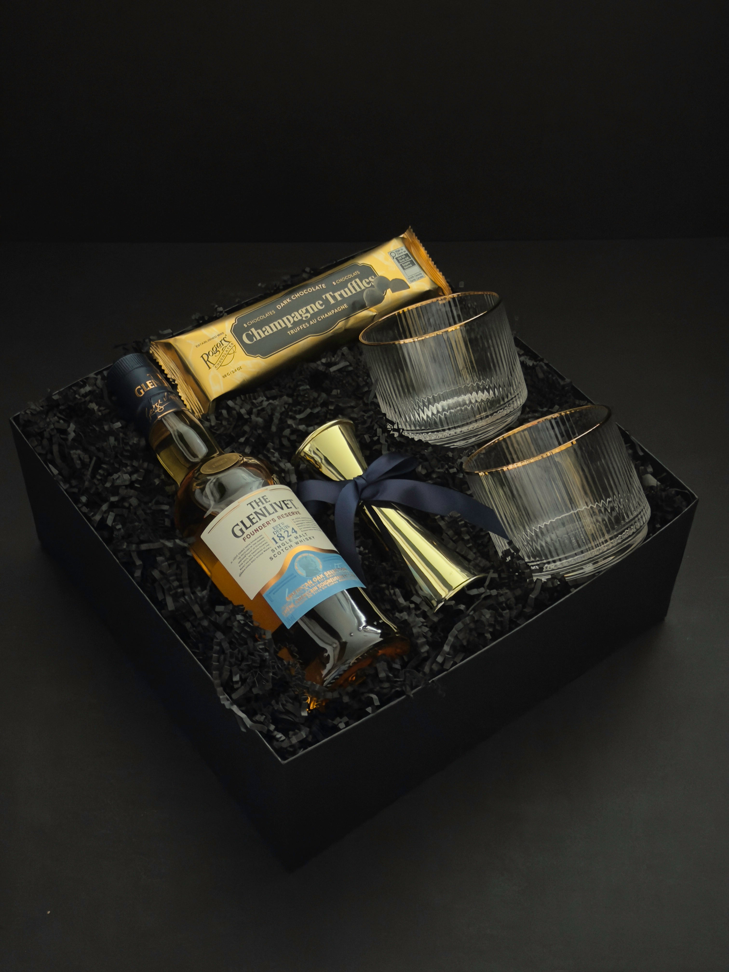 The Belgravia luxury whisky gift basket by Muse Baskets and Co. featuring The Glenlivet Founder's Reserve 1824 Single Malt Scotch Whisky, Viski Meridian gold-rimmed crystal lowball tumblers, a gold shot jigger tied with a navy blue satin ribbon, and Roger's Chocolates Dark Chocolate Champagne Truffles, presented in a signature black gift box with black crinkle fill, Vancouver, BC.