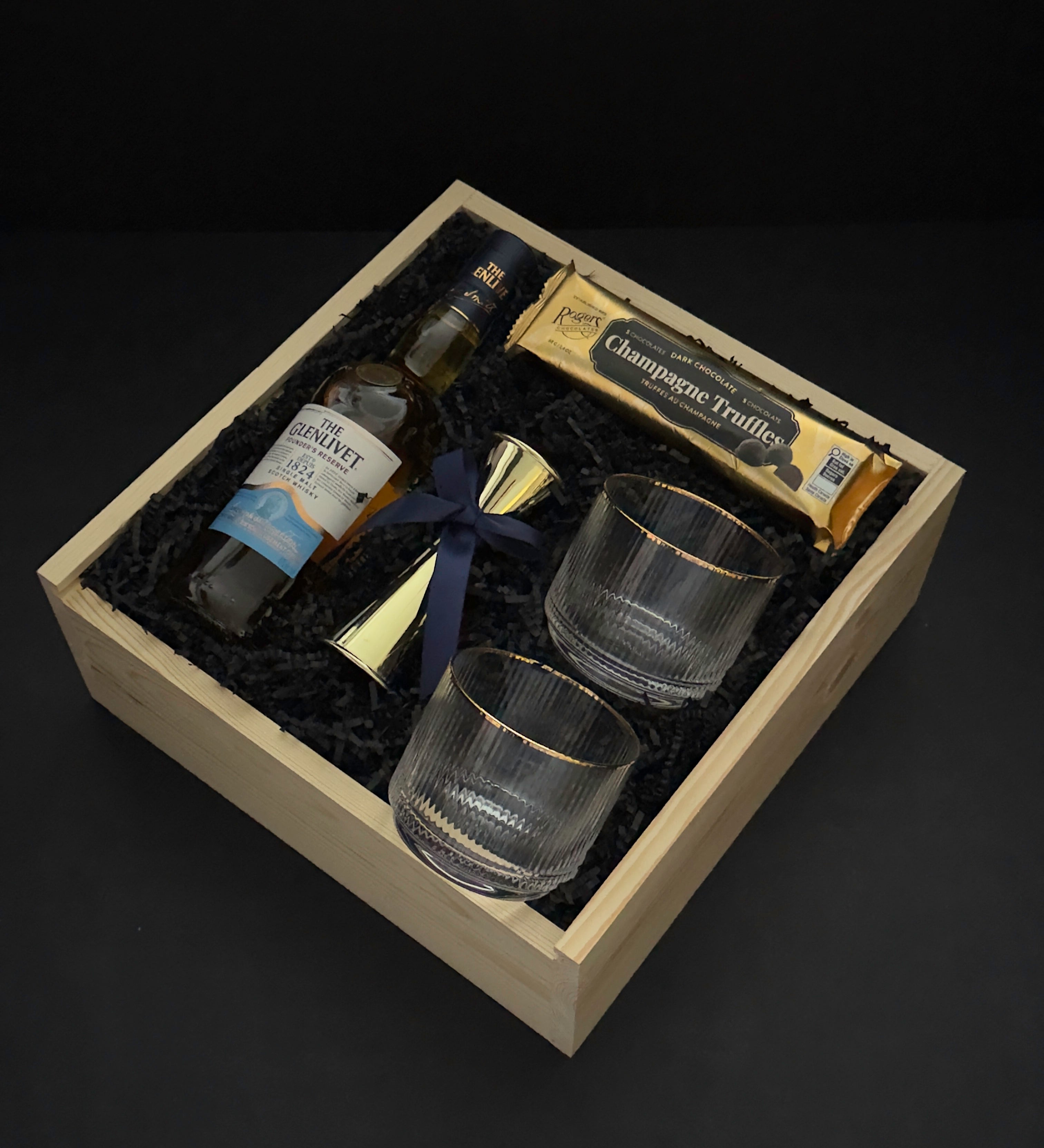 The Belgravia executive gift basket by Muse Baskets and Co. featuring The Glenlivet Founder's Reserve Single Malt Scotch, two Viski Meridian gold-rimmed crystal lowball tumblers, a gold shot jigger with navy blue ribbon, and Roger's Chocolates Dark Chocolate Champagne Truffles, arranged in a signature locally sourced wood gift box with black crinkle fill, realtor closing gift and corporate gift, Vancouver, BC.
