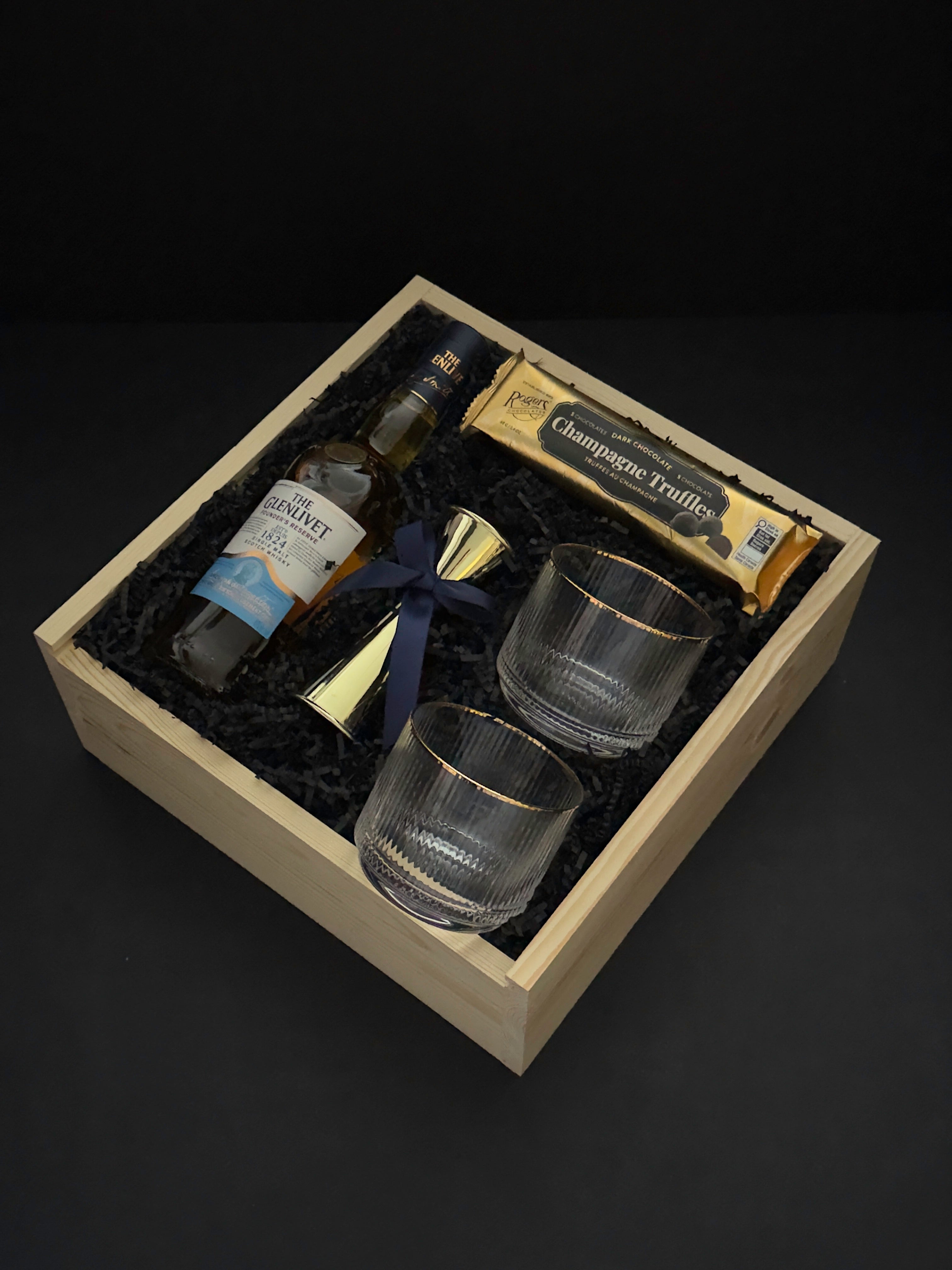 The Belgravia executive gift basket by Muse Baskets and Co. featuring The Glenlivet Founder's Reserve Single Malt Scotch, two Viski Meridian gold-rimmed crystal lowball tumblers, a gold shot jigger with navy blue ribbon, and Roger's Chocolates Dark Chocolate Champagne Truffles, arranged in a signature locally sourced wood gift box with black crinkle fill, realtor closing gift and corporate gift, Vancouver, BC.
