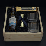 The Belgravia luxury corporate gift basket by Muse Baskets and Co. featuring The Glenlivet Founder's Reserve 1824 Single Malt Scotch Whisky, two Viski Meridian gold-rimmed crystal lowball tumblers, a gold shot jigger tied with a navy satin ribbon, and Roger's Chocolates Dark Chocolate Champagne Truffles, styled in a natural wood gift box with black crinkle fill, possession day gift and executive gift, Vancouver, BC.