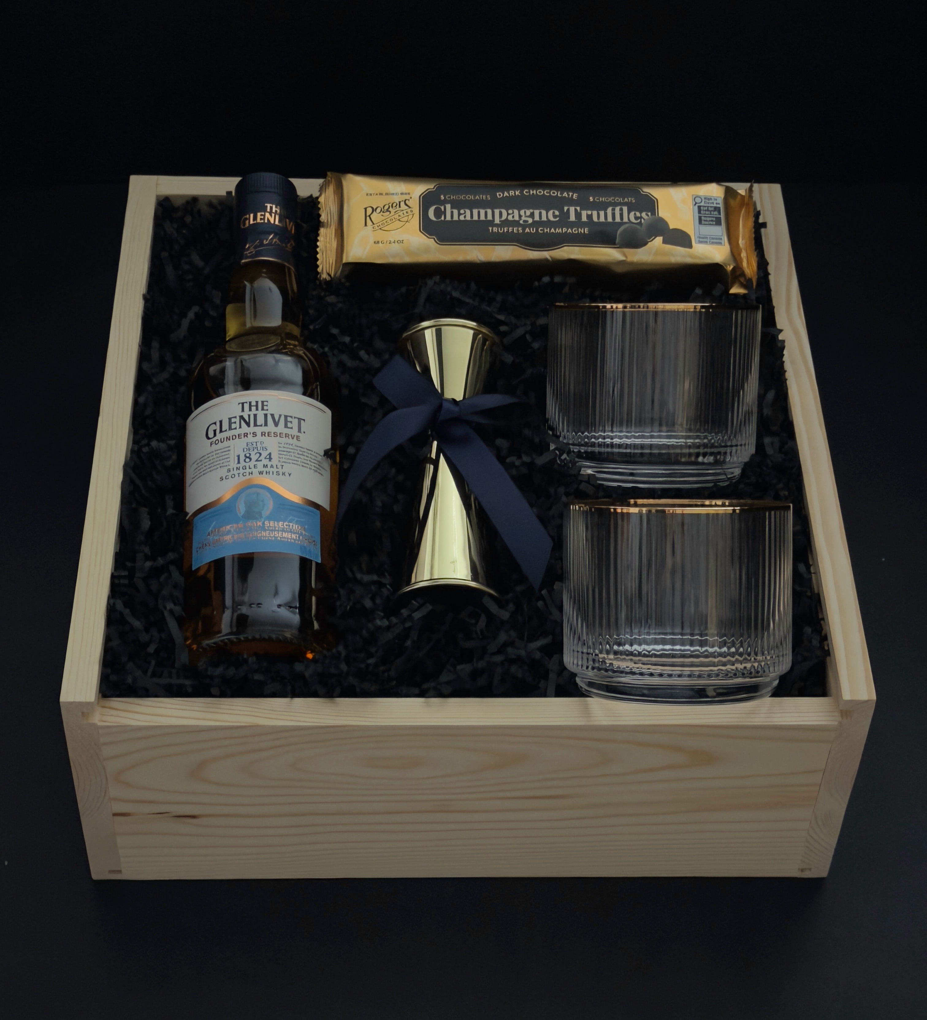 The Belgravia luxury corporate gift basket by Muse Baskets and Co. featuring The Glenlivet Founder's Reserve 1824 Single Malt Scotch Whisky, two Viski Meridian gold-rimmed crystal lowball tumblers, a gold shot jigger tied with a navy satin ribbon, and Roger's Chocolates Dark Chocolate Champagne Truffles, styled in a natural wood gift box with black crinkle fill, possession day gift and executive gift, Vancouver, BC.