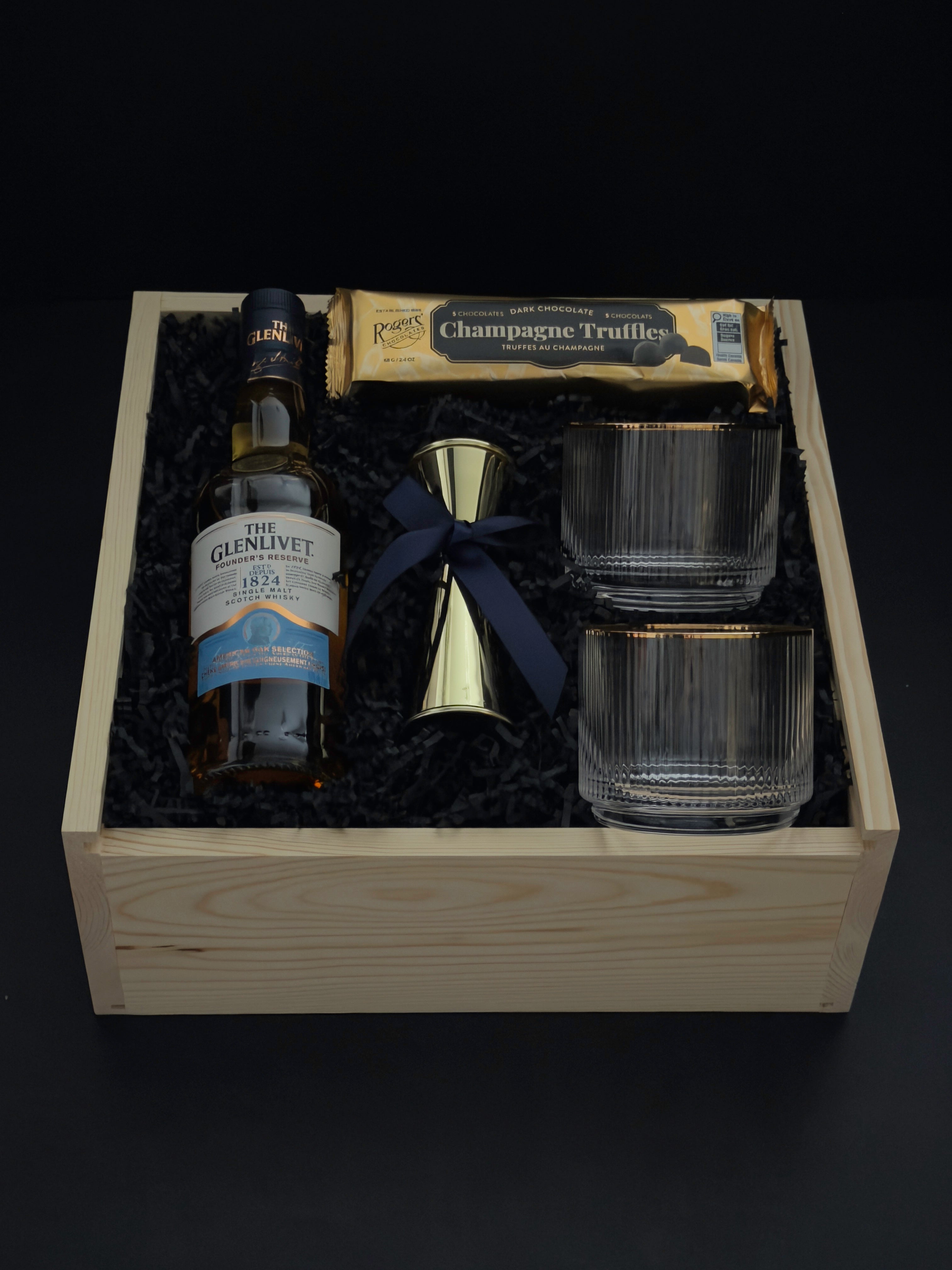 The Belgravia luxury corporate gift basket by Muse Baskets and Co. featuring The Glenlivet Founder's Reserve 1824 Single Malt Scotch Whisky, two Viski Meridian gold-rimmed crystal lowball tumblers, a gold shot jigger tied with a navy satin ribbon, and Roger's Chocolates Dark Chocolate Champagne Truffles, styled in a natural wood gift box with black crinkle fill, possession day gift and executive gift, Vancouver, BC.