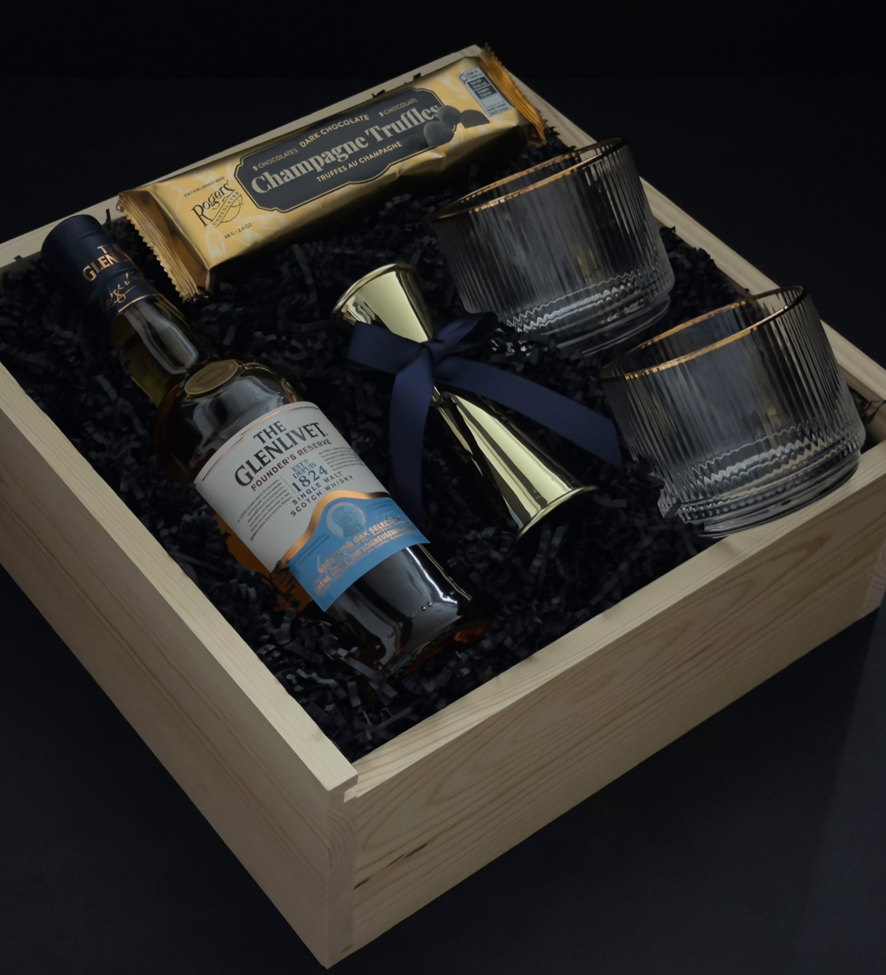 The Belgravia bespoke whisky gift set by Muse Baskets and Co. featuring The Glenlivet Founder's Reserve Single Malt Scotch, Viski Meridian gold-rimmed crystal tumblers, a gold double-sided shot jigger with navy blue bow, and Roger's Chocolates Dark Chocolate Champagne Truffles, presented in a signature locally sourced wood gift box, realtor closing gift and branded corporate gift, Vancouver, BC.