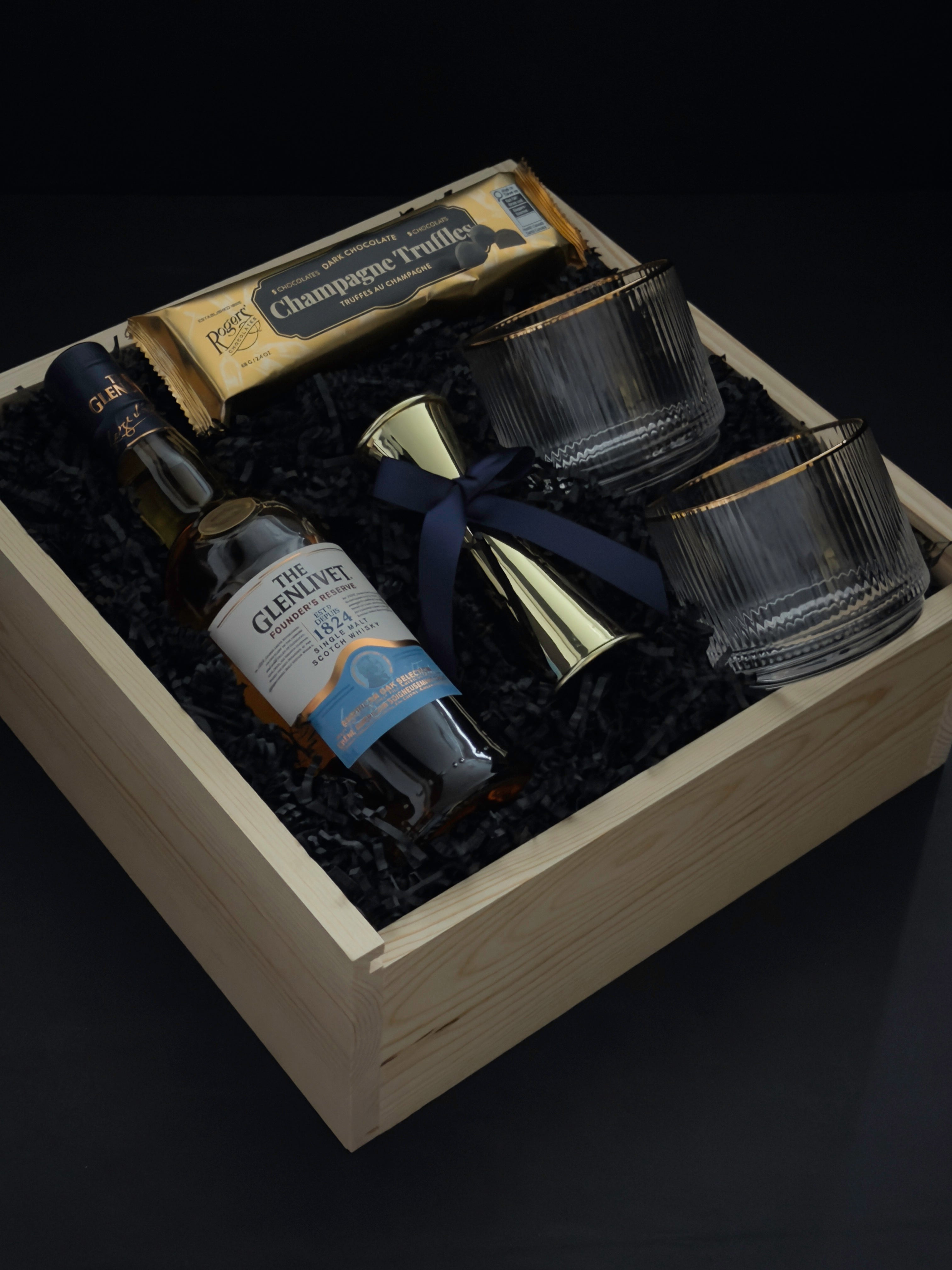 The Belgravia bespoke whisky gift set by Muse Baskets and Co. featuring The Glenlivet Founder's Reserve Single Malt Scotch, Viski Meridian gold-rimmed crystal tumblers, a gold double-sided shot jigger with navy blue bow, and Roger's Chocolates Dark Chocolate Champagne Truffles, presented in a signature locally sourced wood gift box, realtor closing gift and branded corporate gift, Vancouver, BC.