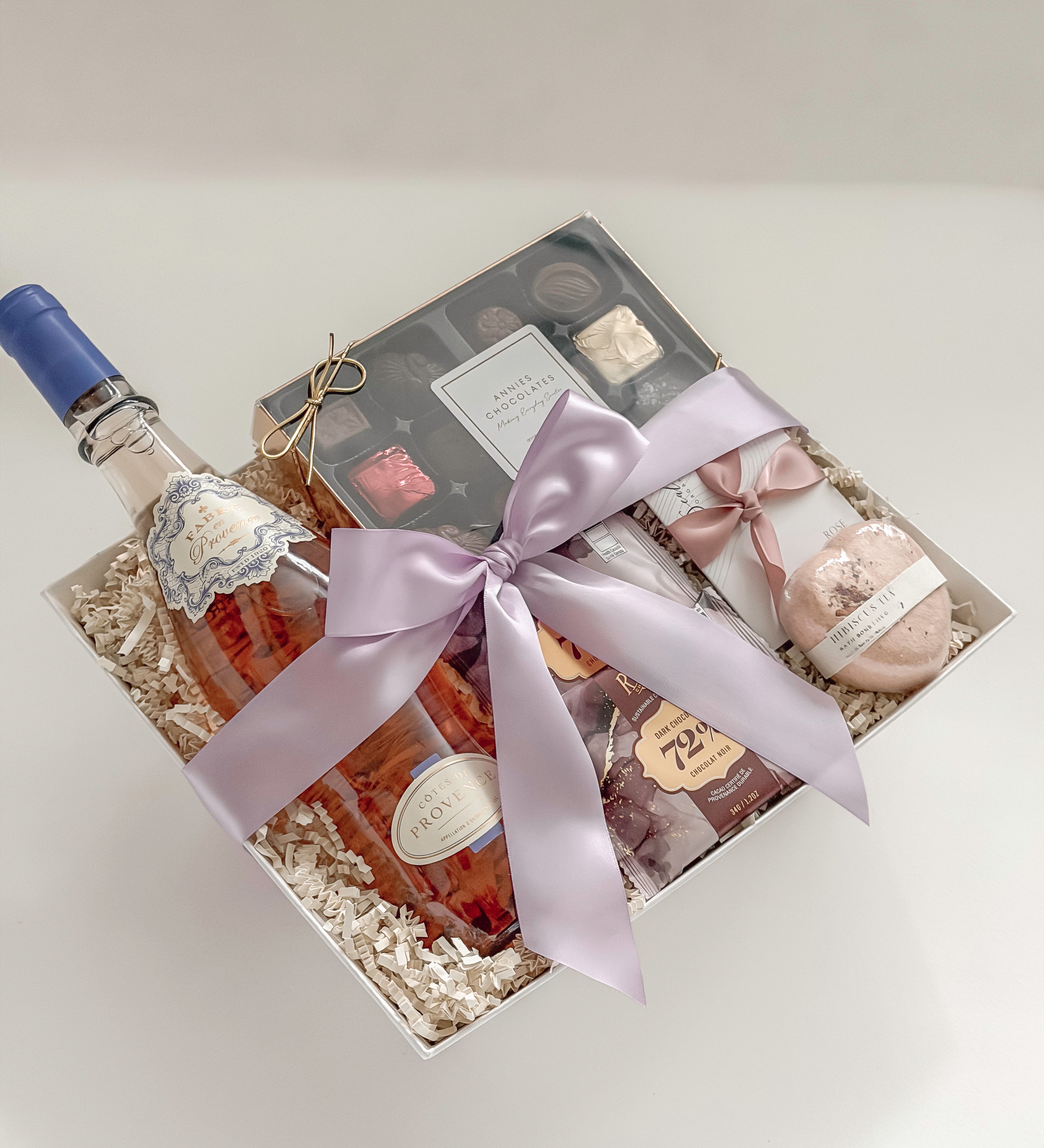 Aurelia celebration gift basket by Muse Baskets and Co. photographed close up, with Côtes de Provence rosé, Annie's assorted chocolates, Roger's 72% dark chocolate bars, and a hibiscus tea heart bath bomb arranged in a white box finished with a light orchid satin bow, a curated gift for her, brides-to-be, and women founders from Canadian makers.