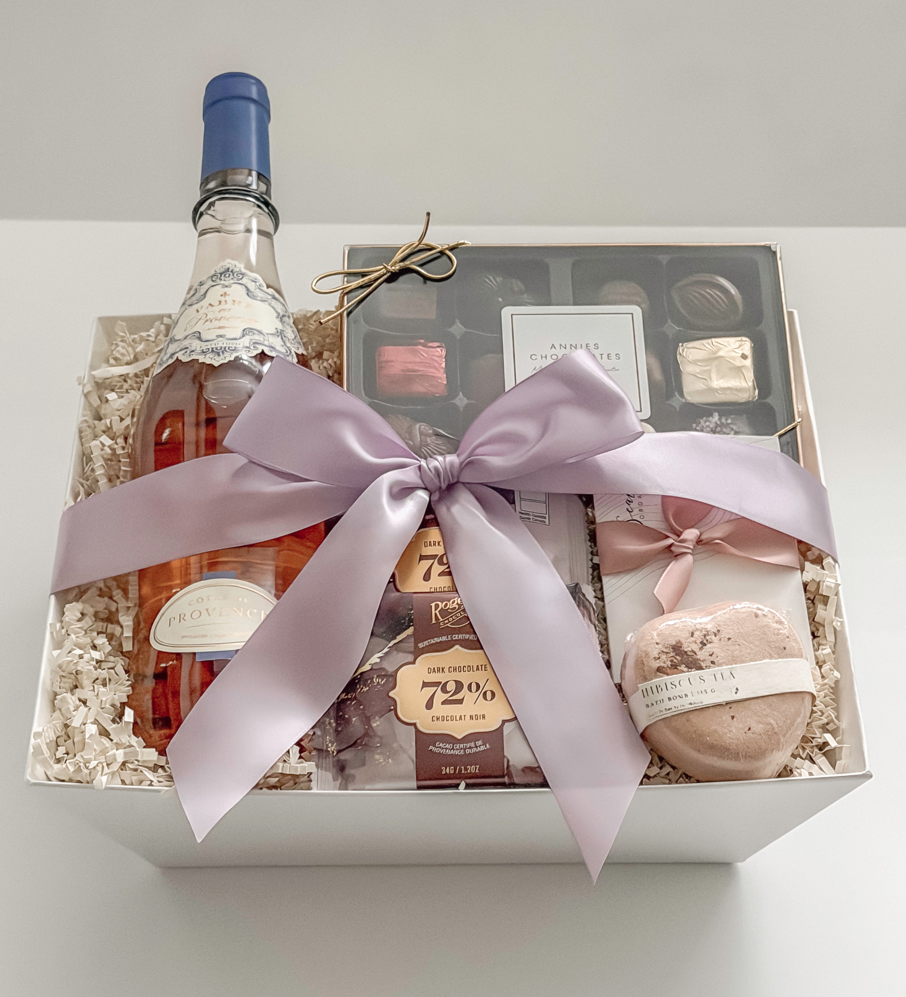 Aurelia gift basket by Muse Baskets and Co. shown from above at an angle, featuring Côtes de Provence rosé by Fabre en Provence, a box of Annie's assorted chocolates, Roger's 72% dark chocolate, a Sealuxe rose face mask, and a hibiscus heart bath bomb in a white box tied with a light orchid bow, a refined bridal shower, birthday, and celebration gift for her.