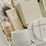 Close-up detail of the Gratitude luxury gift basket by Muse Baskets and Co., a realtor closing and possession day gift, showing the Silentium bergamot, lavender, and musk candle by Vancouver Candle Co., a Laura Slack artisan chocolate bar in gold Chiyogami paper, white chocolate covered pretzels, Home matches in a glass apothecary jar, Handfuel everything bagel cashews, a white stoneware tea cup, and gold-veined marble coasters.