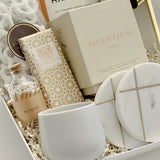 Close-up detail of the Gratitude luxury gift basket by Muse Baskets and Co., a realtor closing and possession day gift, showing the Silentium bergamot, lavender, and musk candle by Vancouver Candle Co., a Laura Slack artisan chocolate bar in gold Chiyogami paper, white chocolate covered pretzels, Home matches in a glass apothecary jar, Handfuel everything bagel cashews, a white stoneware tea cup, and gold-veined marble coasters.