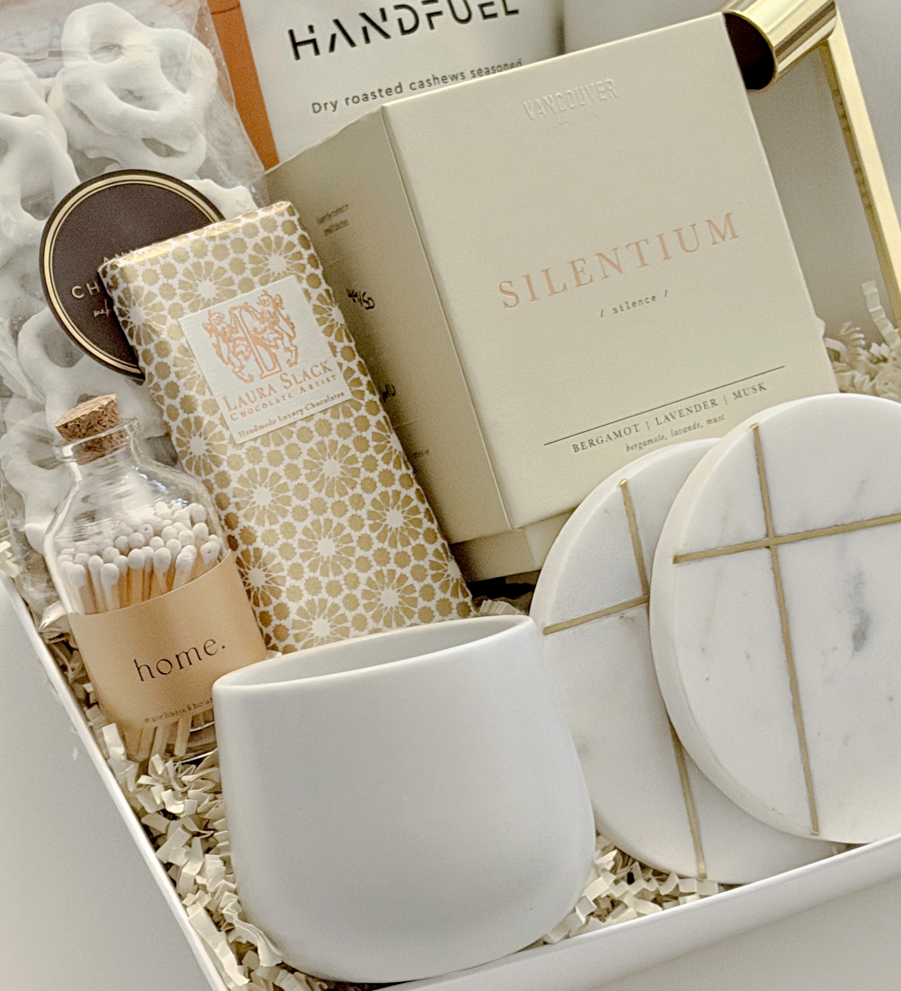 Close-up detail of the Gratitude luxury gift basket by Muse Baskets and Co., a realtor closing and possession day gift, showing the Silentium bergamot, lavender, and musk candle by Vancouver Candle Co., a Laura Slack artisan chocolate bar in gold Chiyogami paper, white chocolate covered pretzels, Home matches in a glass apothecary jar, Handfuel everything bagel cashews, a white stoneware tea cup, and gold-veined marble coasters.