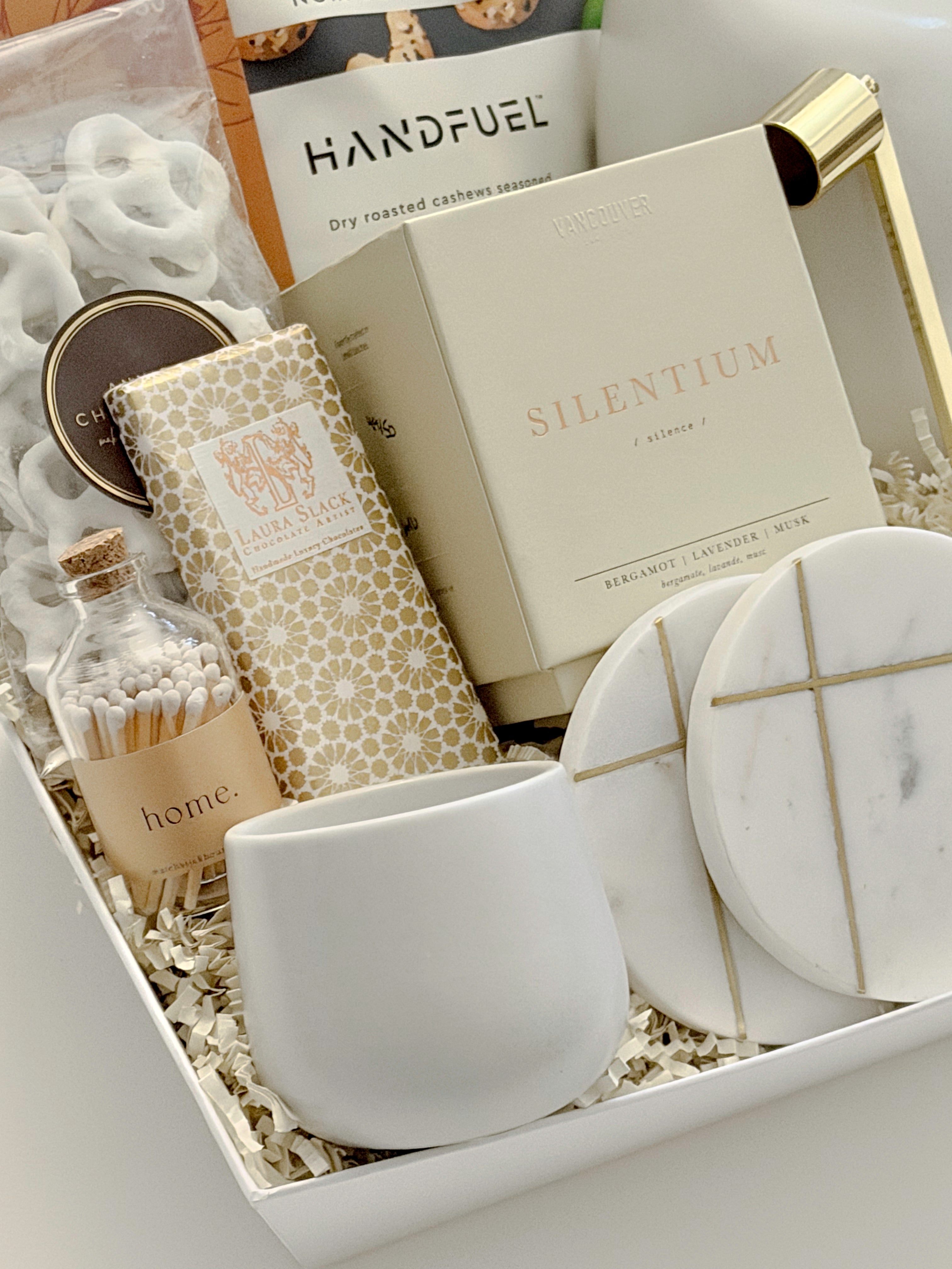 Close-up detail of the Gratitude luxury gift basket by Muse Baskets and Co., a realtor closing and possession day gift, showing the Silentium bergamot, lavender, and musk candle by Vancouver Candle Co., a Laura Slack artisan chocolate bar in gold Chiyogami paper, white chocolate covered pretzels, Home matches in a glass apothecary jar, Handfuel everything bagel cashews, a white stoneware tea cup, and gold-veined marble coasters.