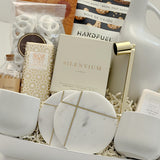 Angled view of the Gratitude gift basket by Muse Baskets and Co., a Canadian artisan realtor closing and homeowner welcome gift, featuring Bush Berry masala chai, Handfuel everything bagel cashews, the Silentium candle by Vancouver Candle Co., a gold candle snuffer, two round marble coasters with gold detailing, white stoneware tea cups, a Laura Slack chocolate bar, and a Fresh Wife vanilla cream bath bomb.