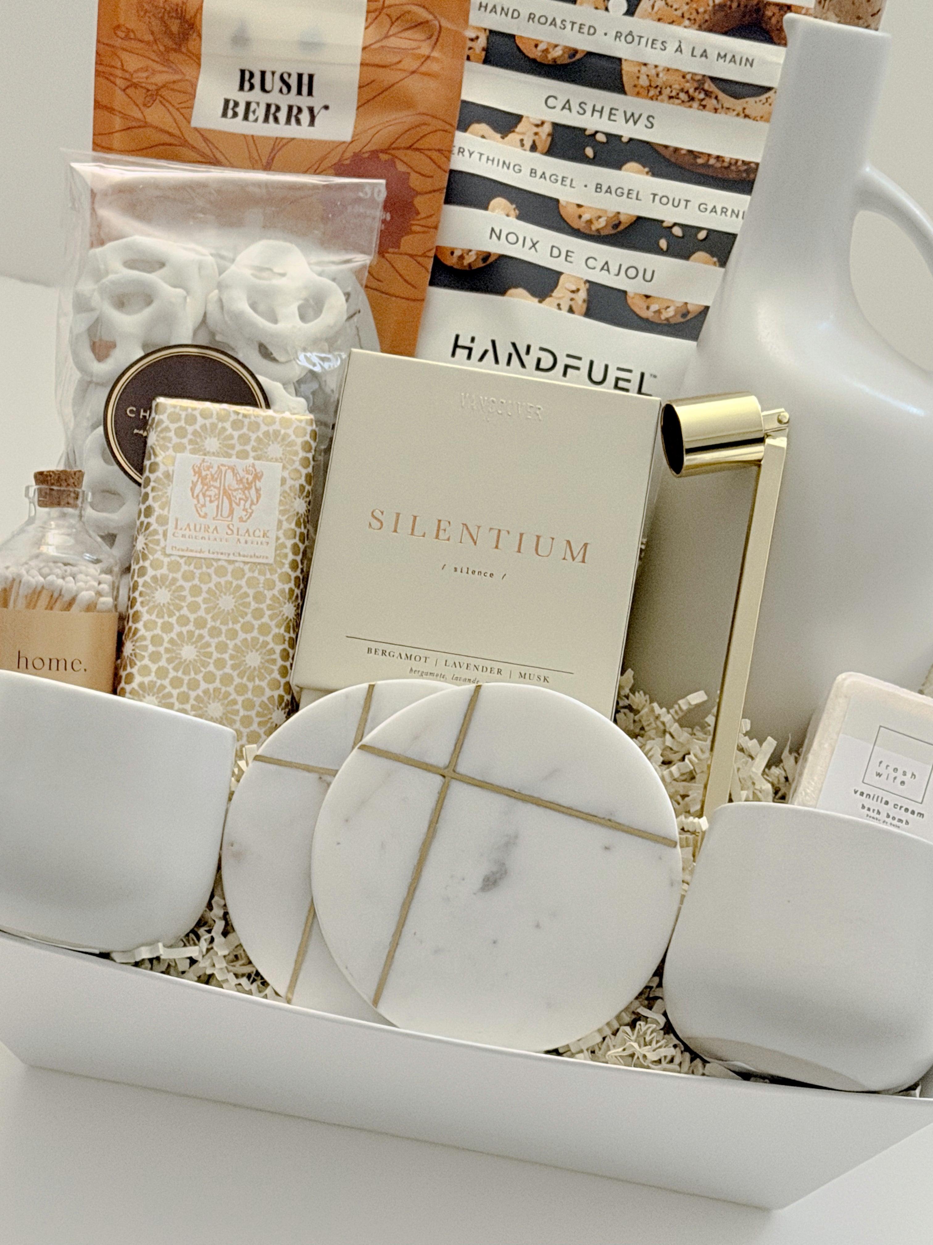 Angled view of the Gratitude gift basket by Muse Baskets and Co., a Canadian artisan realtor closing and homeowner welcome gift, featuring Bush Berry masala chai, Handfuel everything bagel cashews, the Silentium candle by Vancouver Candle Co., a gold candle snuffer, two round marble coasters with gold detailing, white stoneware tea cups, a Laura Slack chocolate bar, and a Fresh Wife vanilla cream bath bomb.