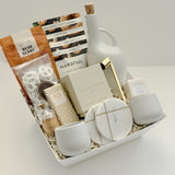 The Gratitude luxury gift basket by Muse Baskets and Co. shown in an open white basket, a possession day and client gift in a neutral white, cream, and gold palette, containing a stoneware olive oil dispenser, Silentium candle, gold snuffer, marble coasters, stoneware tea cups, Bush Berry masala chai, Handfuel cashews, Laura Slack chocolate, white chocolate pretzels, and a Fresh Wife bath bomb on cream paper fill.