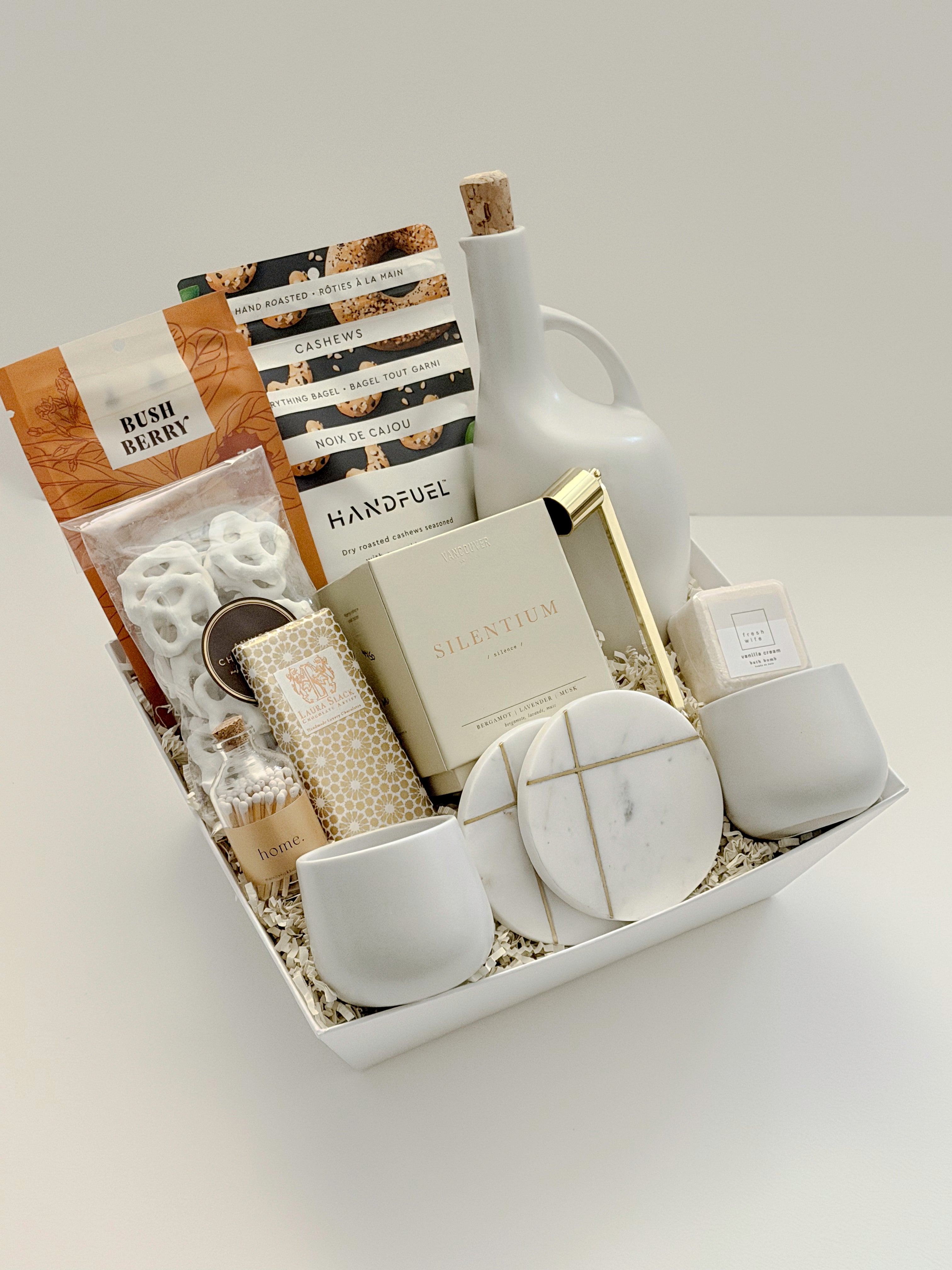 The Gratitude luxury gift basket by Muse Baskets and Co. shown in an open white basket, a possession day and client gift in a neutral white, cream, and gold palette, containing a stoneware olive oil dispenser, Silentium candle, gold snuffer, marble coasters, stoneware tea cups, Bush Berry masala chai, Handfuel cashews, Laura Slack chocolate, white chocolate pretzels, and a Fresh Wife bath bomb on cream paper fill.