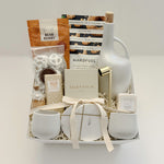 Overhead flat lay of the Gratitude gift basket by Muse Baskets and Co., a Canadian artisan realtor closing, possession day, and corporate gift, finished with a satin ribbon and arranged in a white basket with a stoneware olive oil dispenser, Silentium candle, gold candle snuffer, gold-veined marble coasters, two white stoneware tea cups, Bush Berry masala chai, Handfuel cashews, Laura Slack chocolate, chocolate covered pretzels, and a Fresh Wife vanilla cream bath bomb.
