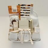 Overhead flat lay of the Gratitude gift basket by Muse Baskets and Co., a Canadian artisan realtor closing, possession day, and corporate gift, finished with a satin ribbon and arranged in a white basket with a stoneware olive oil dispenser, Silentium candle, gold candle snuffer, gold-veined marble coasters, two white stoneware tea cups, Bush Berry masala chai, Handfuel cashews, Laura Slack chocolate, chocolate covered pretzels, and a Fresh Wife vanilla cream bath bomb.