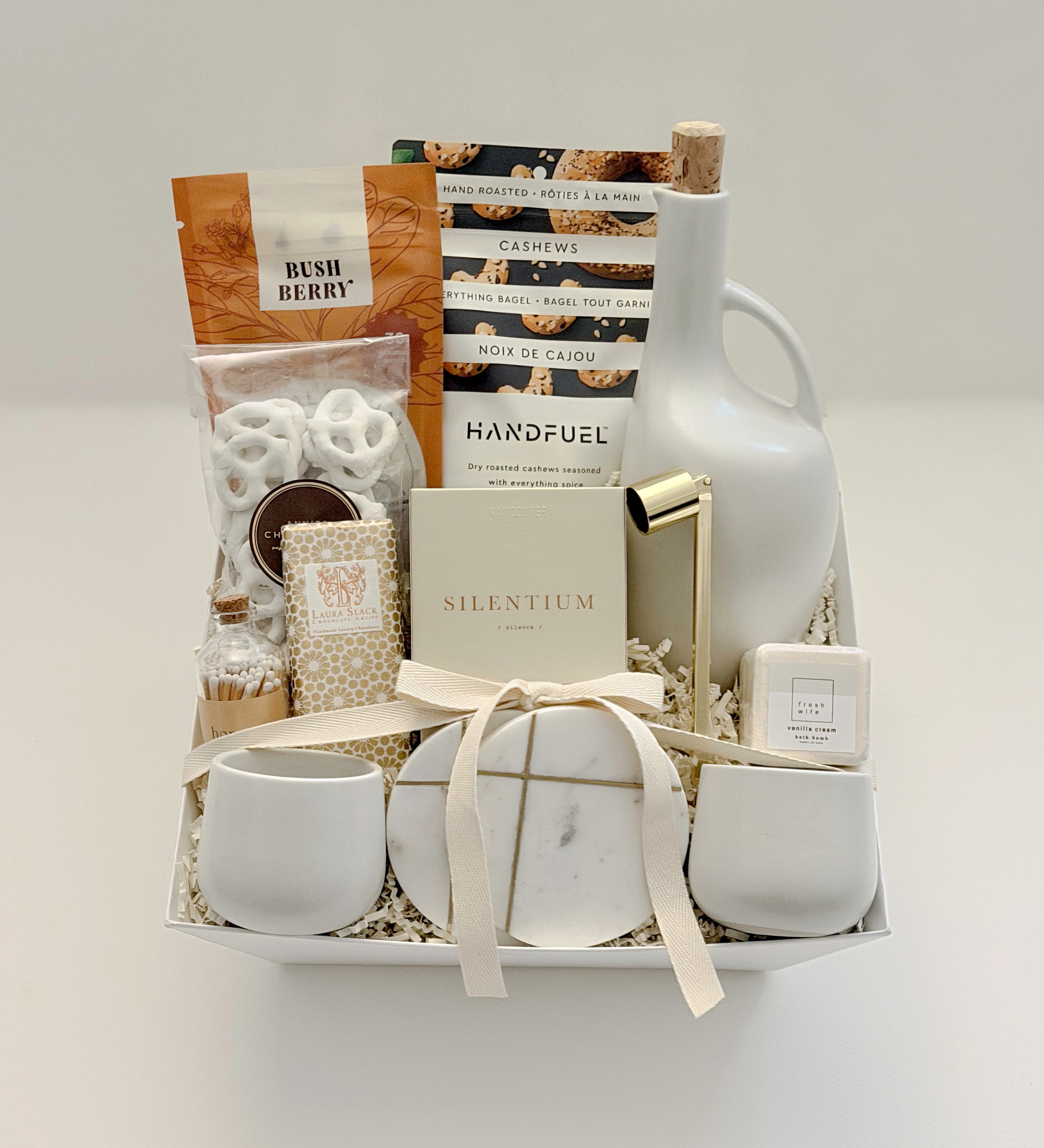 Overhead flat lay of the Gratitude gift basket by Muse Baskets and Co., a Canadian artisan realtor closing, possession day, and corporate gift, finished with a satin ribbon and arranged in a white basket with a stoneware olive oil dispenser, Silentium candle, gold candle snuffer, gold-veined marble coasters, two white stoneware tea cups, Bush Berry masala chai, Handfuel cashews, Laura Slack chocolate, chocolate covered pretzels, and a Fresh Wife vanilla cream bath bomb.