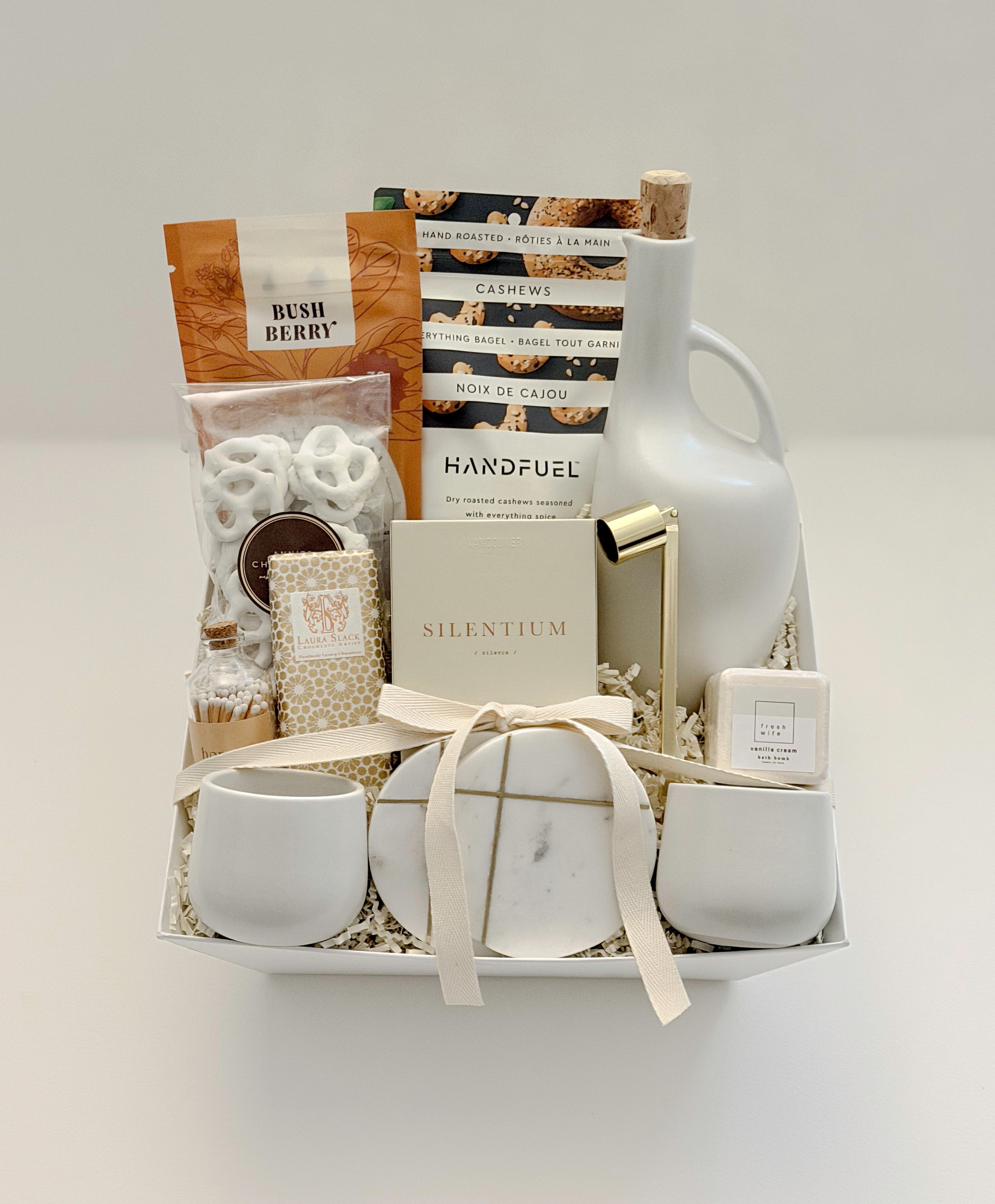 Overhead flat lay of the Gratitude gift basket by Muse Baskets and Co., a Canadian artisan realtor closing, possession day, and corporate gift, finished with a satin ribbon and arranged in a white basket with a stoneware olive oil dispenser, Silentium candle, gold candle snuffer, gold-veined marble coasters, two white stoneware tea cups, Bush Berry masala chai, Handfuel cashews, Laura Slack chocolate, chocolate covered pretzels, and a Fresh Wife vanilla cream bath bomb.
