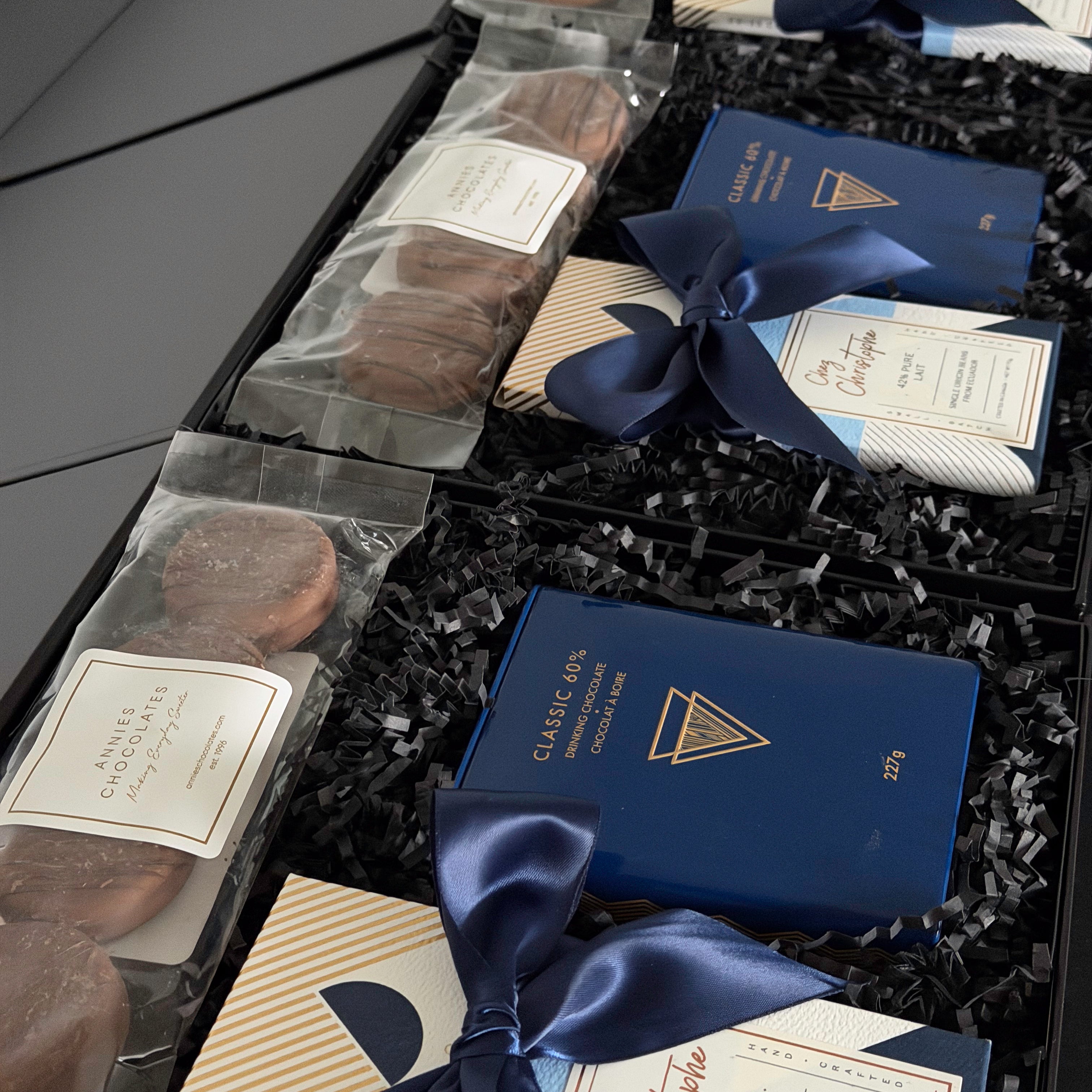 A luxury chocolate gift box filled with assorted dark chocolate cookies in clear bags and boxed chocolates tied with navy blue ribbons, arranged in black crinkle paper.