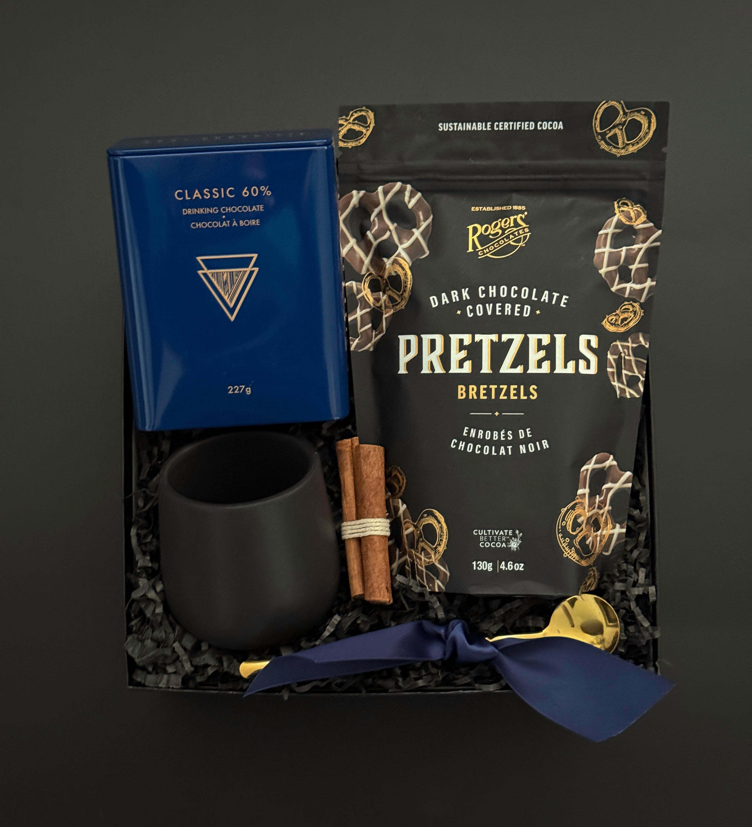 The Midnight Cocoa Gift Box displayed with artisan drinking chocolate, matte black stoneware cup, gold stirring spoon, dark chocolate pretzels, and festive cinnamon accents on a soft winter backdrop.