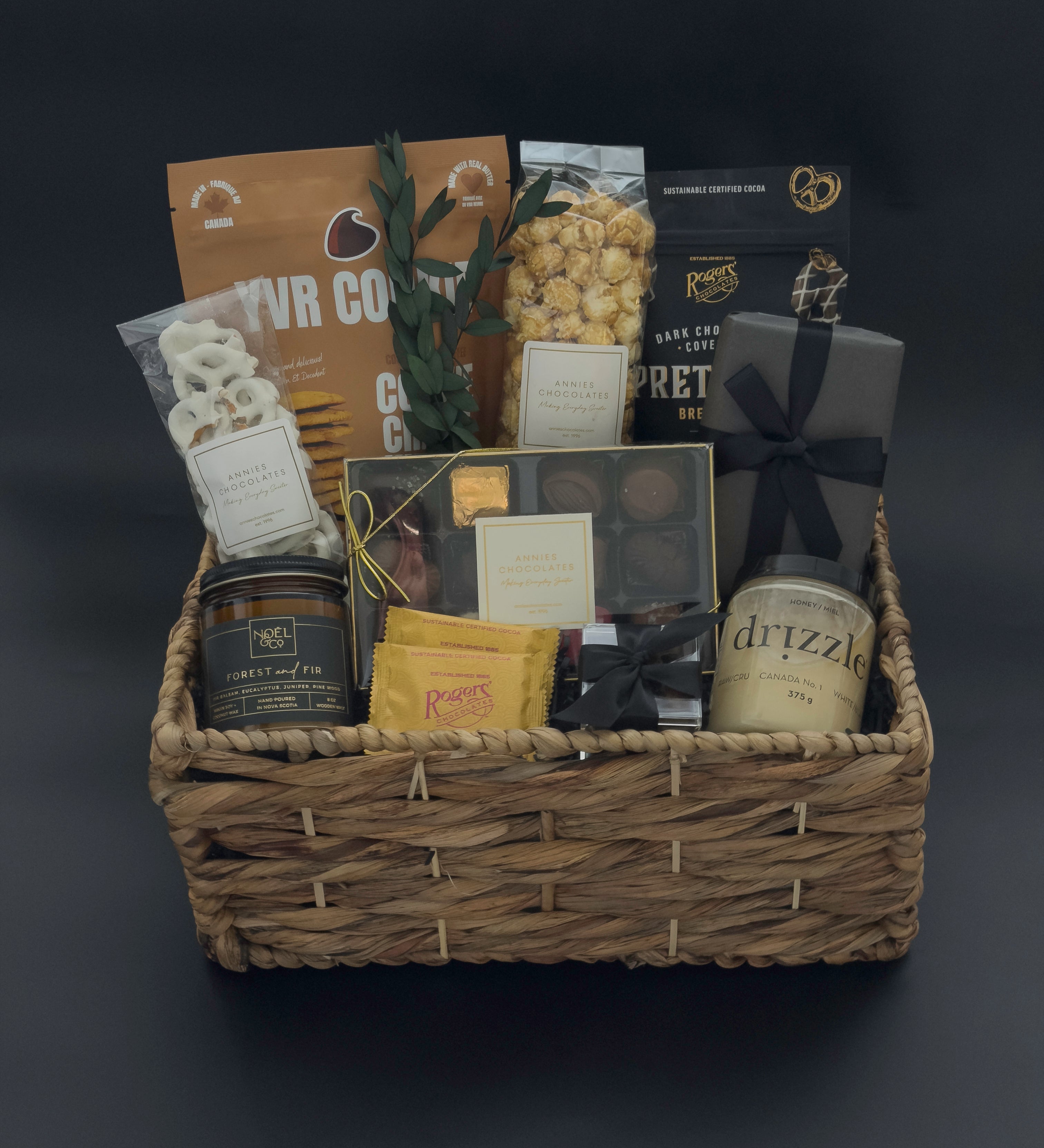 Luxury metropolitan gift basket featuring gourmet chocolates, cookies, raw honey, and forest fir candle arranged in a woven basket with black satin ribbon