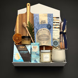 Fireside Provisions by Muse Baskets and Co., a Canadian holiday gift box with Camembert, cashews and chocolate pretzels arranged with seasonal greenery.