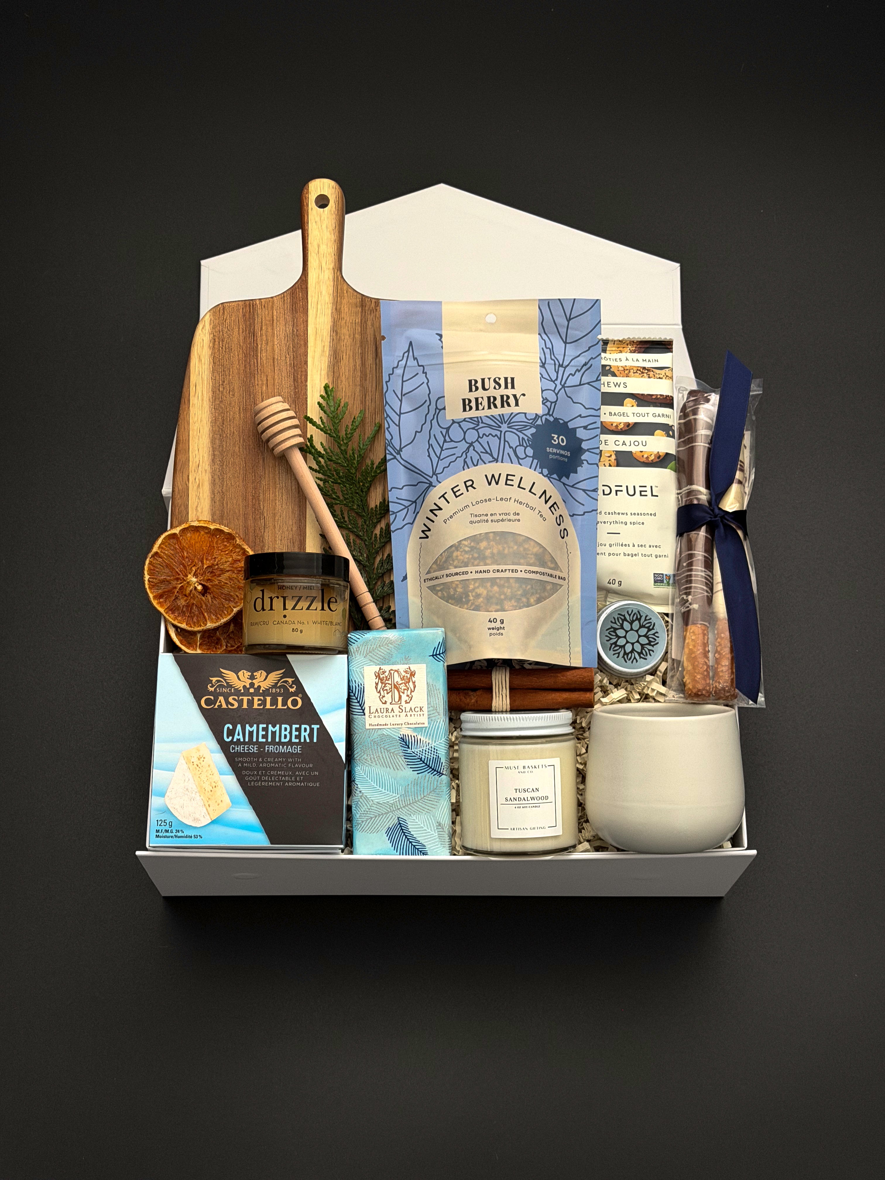 Fireside Provisions by Muse Baskets and Co., a Canadian holiday gift box with Camembert, cashews and chocolate pretzels arranged with seasonal greenery.