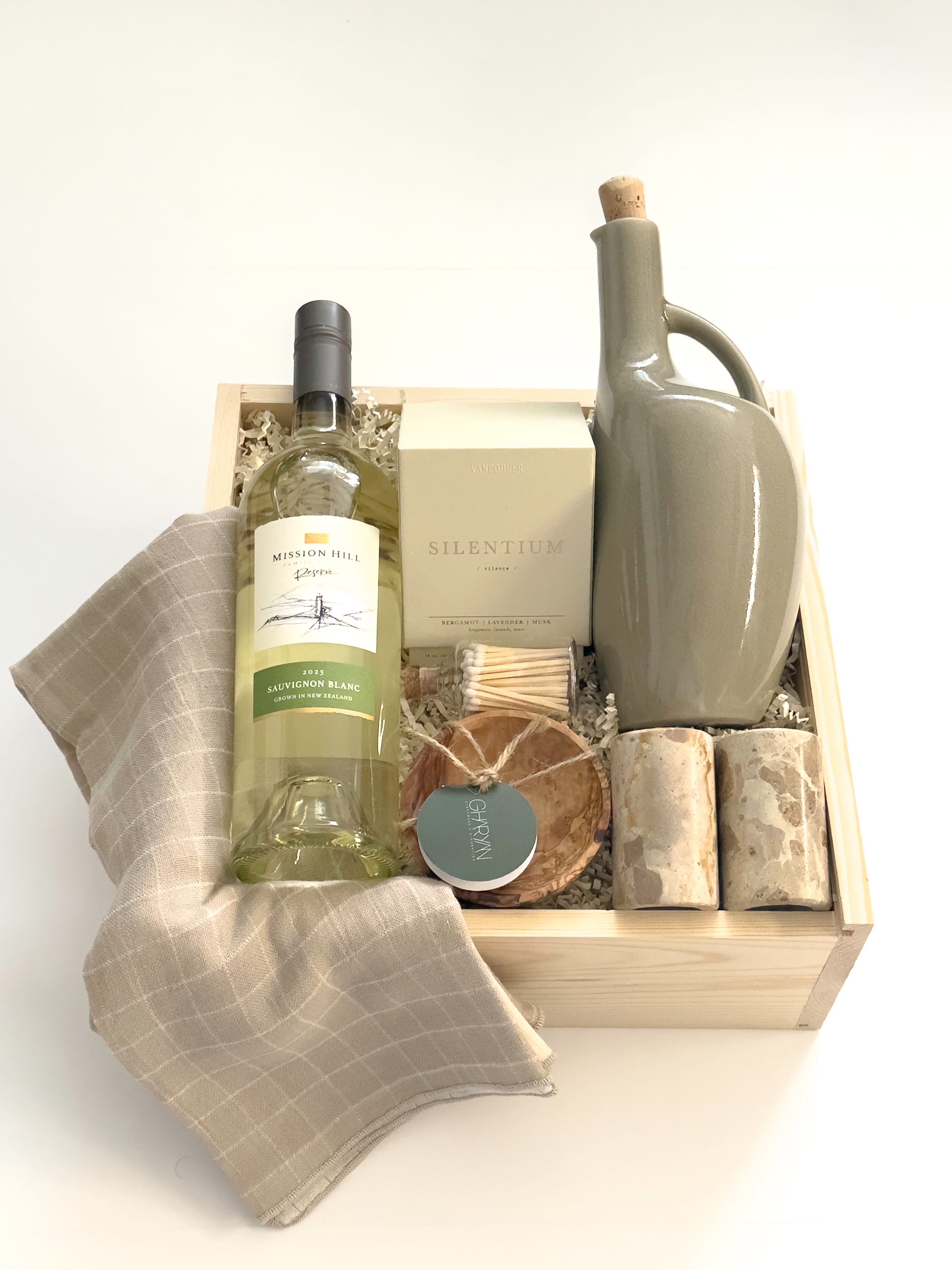The Harvest Host Thanksgiving gift basket by Muse Baskets and Co. featuring Mission Hill Sauvignon Blanc, a Vancouver Candle Co. candle, matches, stoneware olive oil dispenser, marble salt and pepper shakers, olive wood ramekin and grey dishtowels in a wood gift box.