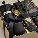 Close-up detail of the Oro Nero luxury coffee gift box by Muse Baskets and Co., showing a matte black Grosche Moka pot, gold coffee scoop, black stoneware cup, So charcoal cleansing bar, Laura Slack artisan chocolate, and a James Street Co. teakwood and tobacco candle, styled with eucalyptus and a black satin ribbon. A refined client gift, corporate gift, and birthday gift