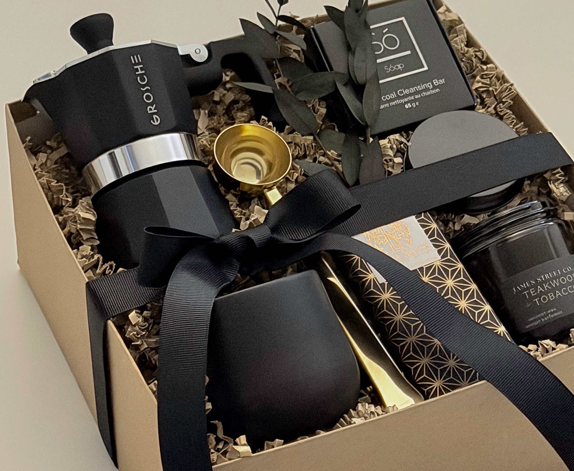 Close-up detail of the Oro Nero luxury coffee gift box by Muse Baskets and Co., showing a matte black Grosche Moka pot, gold coffee scoop, black stoneware cup, So charcoal cleansing bar, Laura Slack artisan chocolate, and a James Street Co. teakwood and tobacco candle, styled with eucalyptus and a black satin ribbon. A refined client gift, corporate gift, and birthday gift
