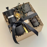 Oro Nero luxury coffee gift box by Muse Baskets and Co. shown at an angle, featuring a Grosche Moka pot, gold coffee scoop, handmade black stoneware cup, So charcoal cleansing bar, Laura Slack chocolate, and a James Street Co. teakwood and tobacco candle, arranged in a kraft box with eucalyptus and a black satin bow. A Canadian artisan client gift and corporate gift.
