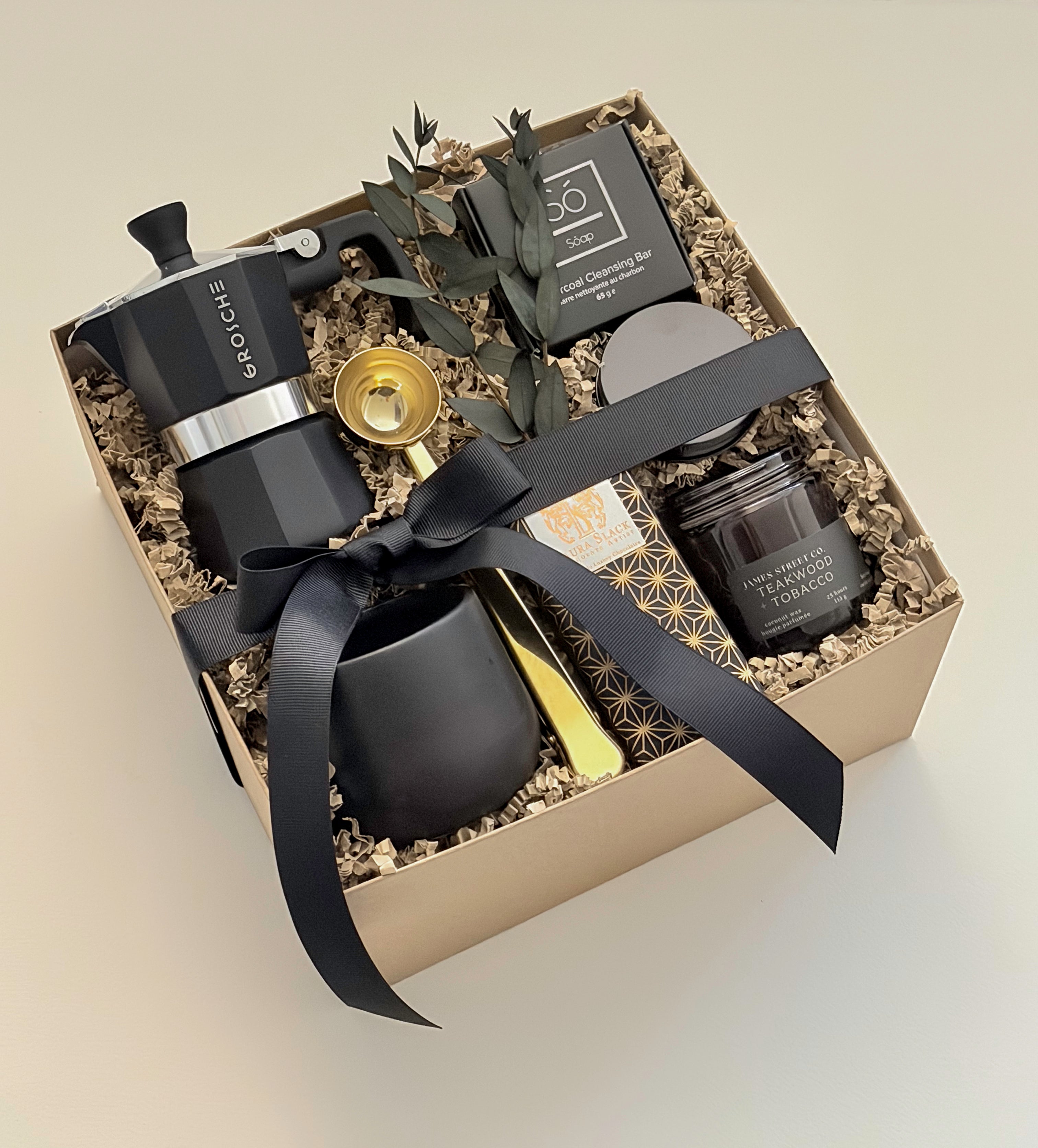 Oro Nero luxury coffee gift box by Muse Baskets and Co. shown at an angle, featuring a Grosche Moka pot, gold coffee scoop, handmade black stoneware cup, So charcoal cleansing bar, Laura Slack chocolate, and a James Street Co. teakwood and tobacco candle, arranged in a kraft box with eucalyptus and a black satin bow. A Canadian artisan client gift and corporate gift.