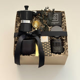 Overhead flat lay of the Oro Nero coffee gift box by Muse Baskets and Co., with a matte black Grosche Moka pot, gold coffee scoop, black stoneware cup, So charcoal cleansing bar, Laura Slack artisan chocolate, and a James Street Co. teakwood and tobacco candle, set in a kraft box with eucalyptus and black ribbon. A luxury client gift and corporate gift with Canada-wide delivery.