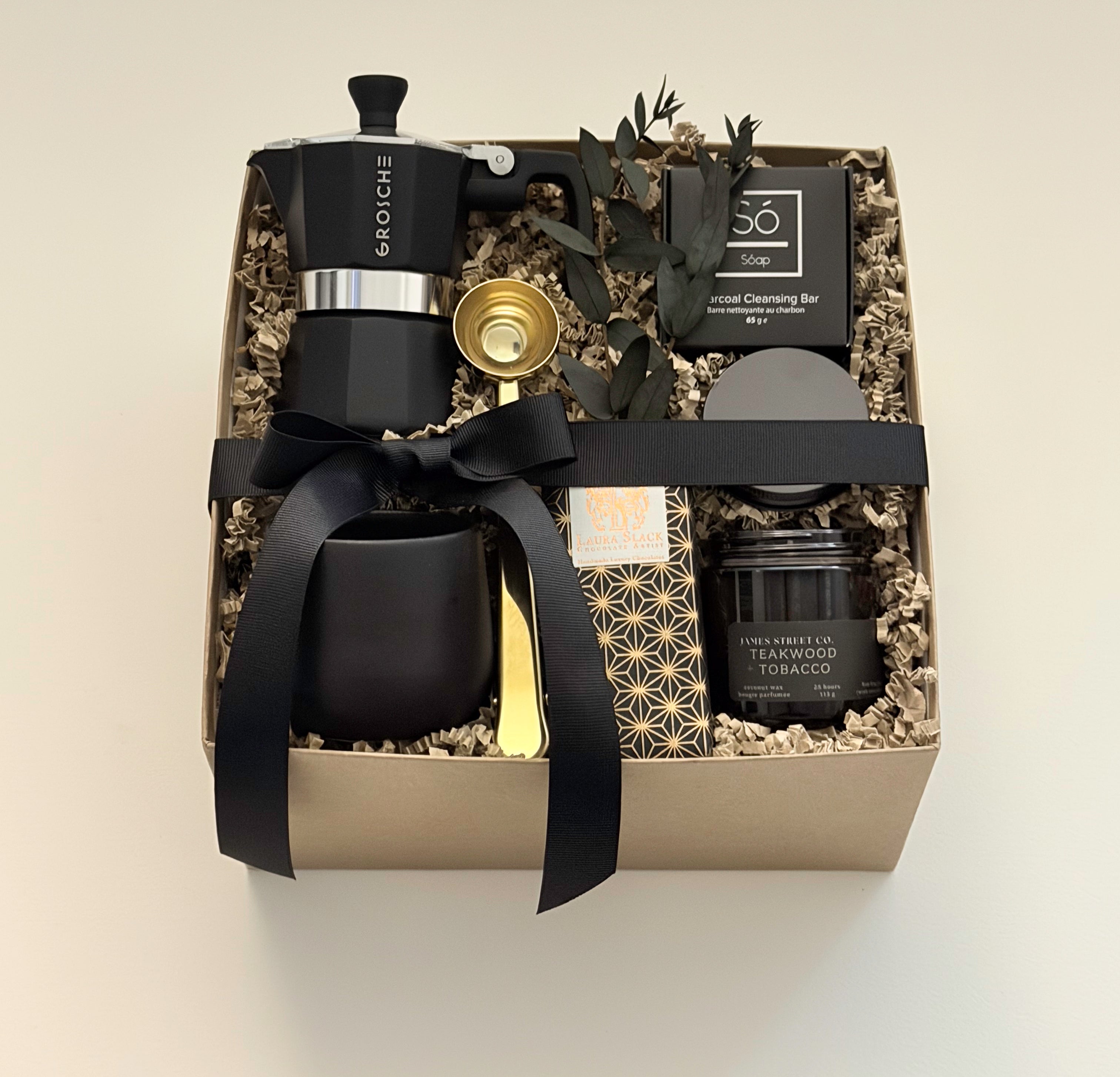 Overhead flat lay of the Oro Nero coffee gift box by Muse Baskets and Co., with a matte black Grosche Moka pot, gold coffee scoop, black stoneware cup, So charcoal cleansing bar, Laura Slack artisan chocolate, and a James Street Co. teakwood and tobacco candle, set in a kraft box with eucalyptus and black ribbon. A luxury client gift and corporate gift with Canada-wide delivery.