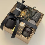 Oro Nero luxury gift box by Muse Baskets and Co. viewed from above at an angle, containing a Grosche stovetop Moka pot, gold coffee scoop, black stoneware cup, So charcoal cleansing bar, Laura Slack chocolate, and a James Street Co. teakwood and tobacco candle, styled with eucalyptus and a black satin ribbon. A bespoke corporate gift and birthday gift for the coffee lover.