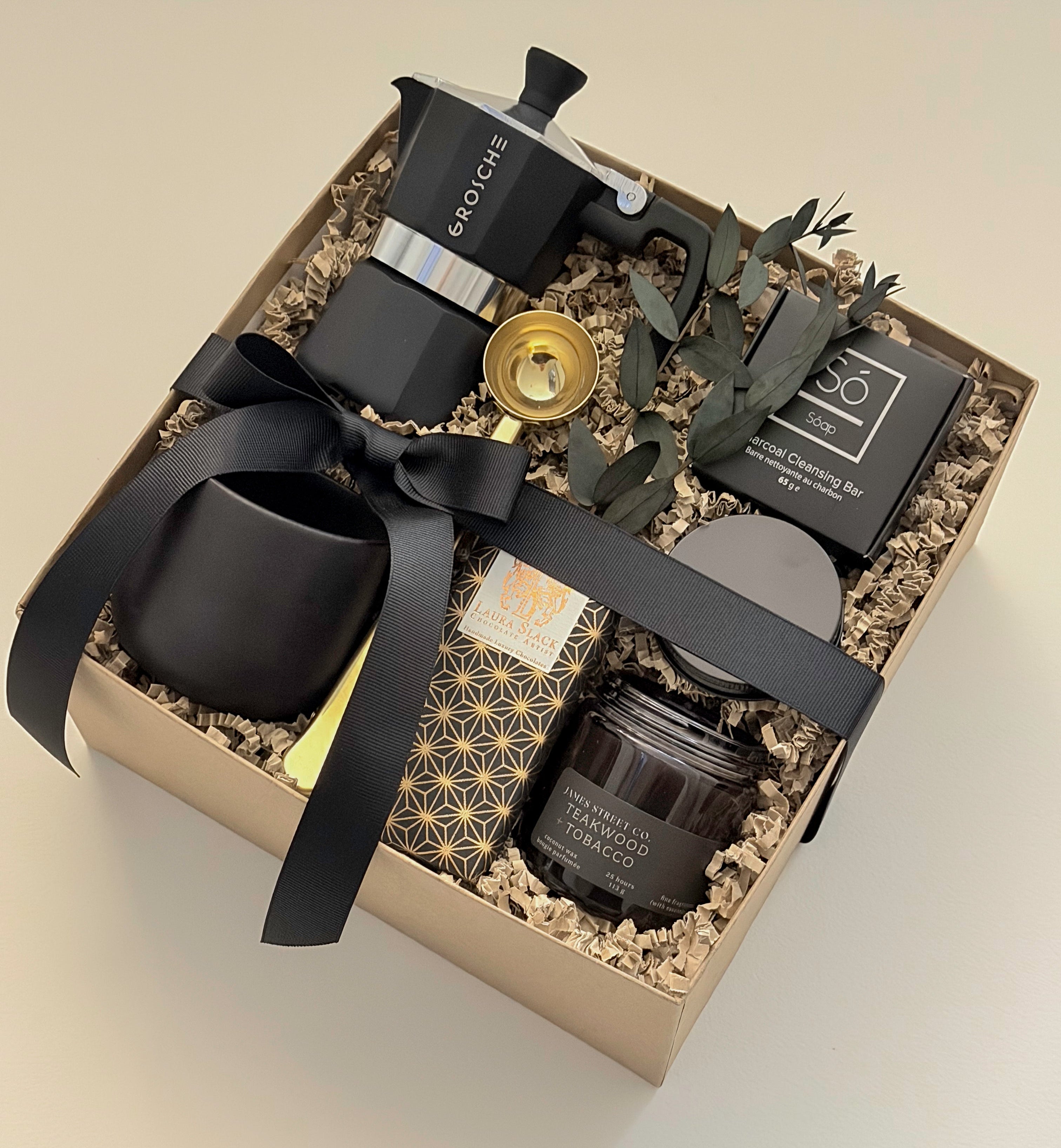 Oro Nero luxury gift box by Muse Baskets and Co. viewed from above at an angle, containing a Grosche stovetop Moka pot, gold coffee scoop, black stoneware cup, So charcoal cleansing bar, Laura Slack chocolate, and a James Street Co. teakwood and tobacco candle, styled with eucalyptus and a black satin ribbon. A bespoke corporate gift and birthday gift for the coffee lover.