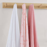 Three swaddles in white, pink, and red hanging on a wooden rod against a light background.