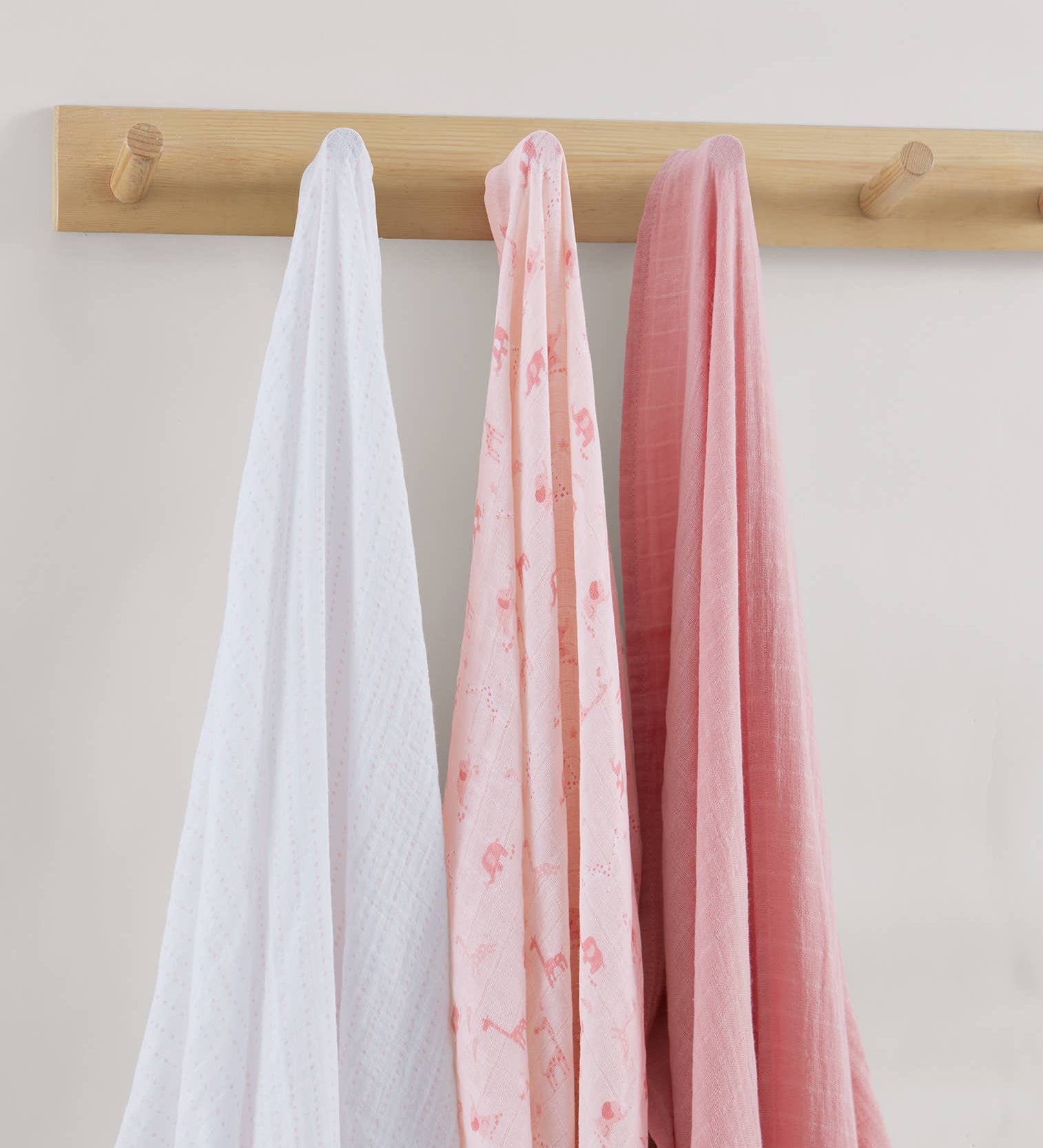 Three swaddles in white, pink, and red hanging on a wooden rod against a light background.